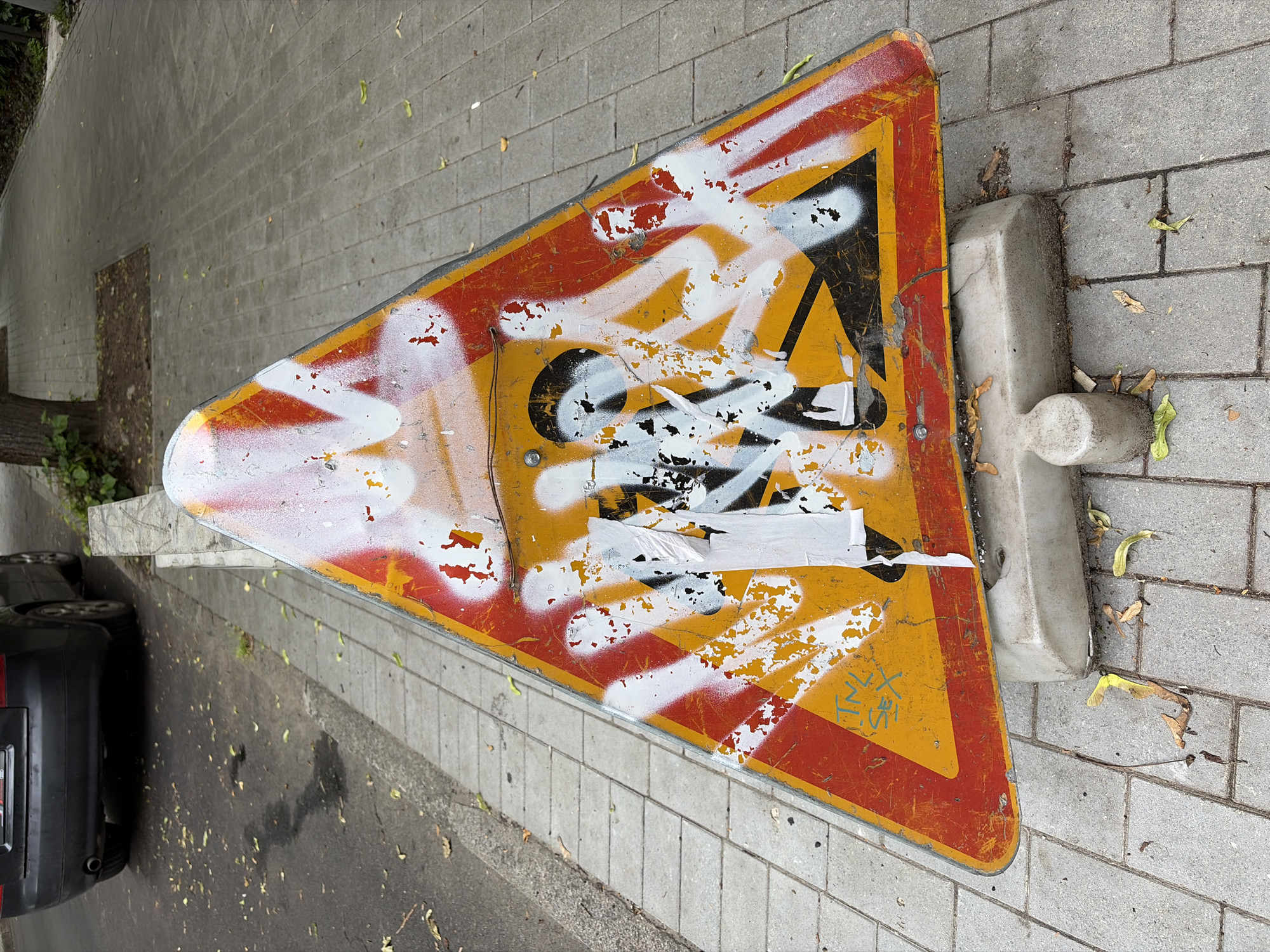A close-up, eye-level shot shows a heavily graffitied triangular road sign on a paved sidewalk. The sign is yellow with red border and depicts silhouetted figures engaged in construction work. White and black spray paint obscures much of the original image. A torn piece of white paper is adhered to the sign. The sign rests on a weathered concrete base, which is placed on a sidewalk made of interlocking gray bricks. Fallen leaves are scattered on the sidewalk and around the base of the sign. To the left of the sidewalk, a portion of a dark-colored car is visible on an asphalt road, with some wet patches and scattered leaves on the road surface. In the background, a paved path continues, bordered by greenery and a tree. The overall impression is one of urban decay and neglect.