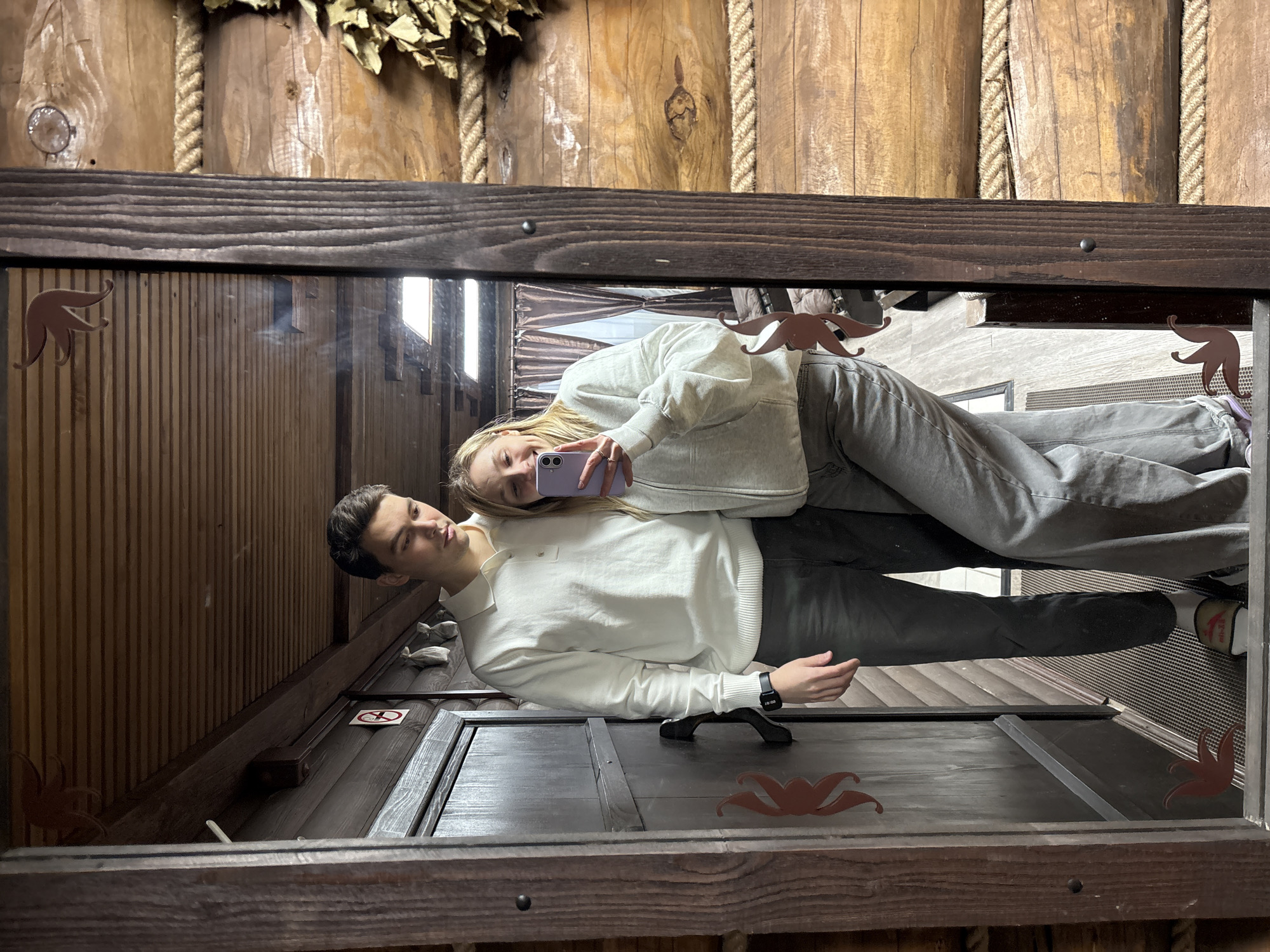 A mirror reflects a young couple in Prikamskii, Russia. The man is wearing a white sweater and grey pants, while the woman is wearing a light-colored hoodie and grey pants, holding a purple cell phone. They are positioned horizontally in the mirror's reflection. The mirror frame is wooden, with decorative leaf-like shapes. The background includes wood paneling, rope details, and bundled dry leaves. The floor visible in the reflection appears to be a dark grey or black color.