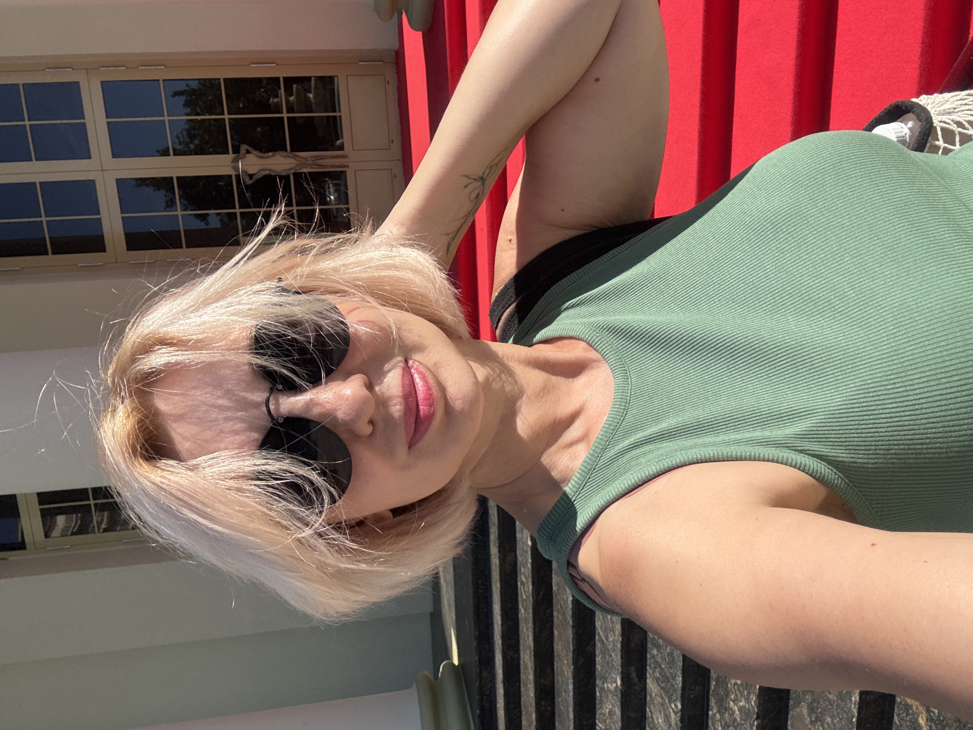 A woman with blonde hair and sunglasses is taking a selfie. She is wearing a green tank top. The background shows a building with large double doors and red carpeted stairs. Based on the context of the question regarding Sochi, Russia, this scene is likely located at the Winter Theatre in Sochi.