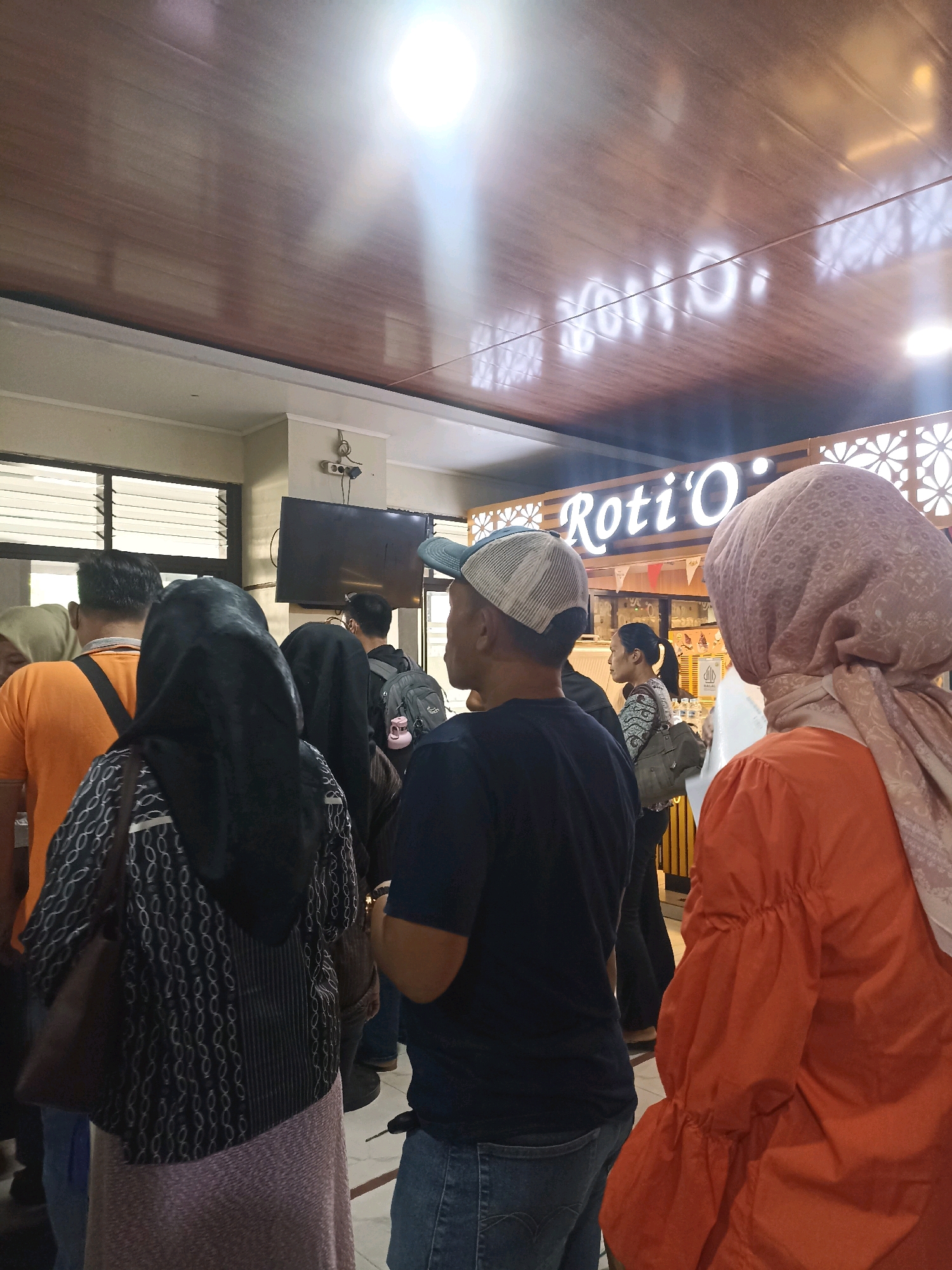 The image shows a queue of people in what appears to be a food court or a shop in Bandar Lampung, Indonesia. The signage in the background reads "Roti O", a popular Indonesian bakery chain. The ceiling is made of dark wood-effect panels, and the lighting is bright, reflecting off the polished surfaces. Several individuals are visible, mostly from the back, dressed in casual attire. Some are wearing headscarves, suggesting a predominantly Muslim population in the area. The overall atmosphere appears to be one of everyday activity and commerce.