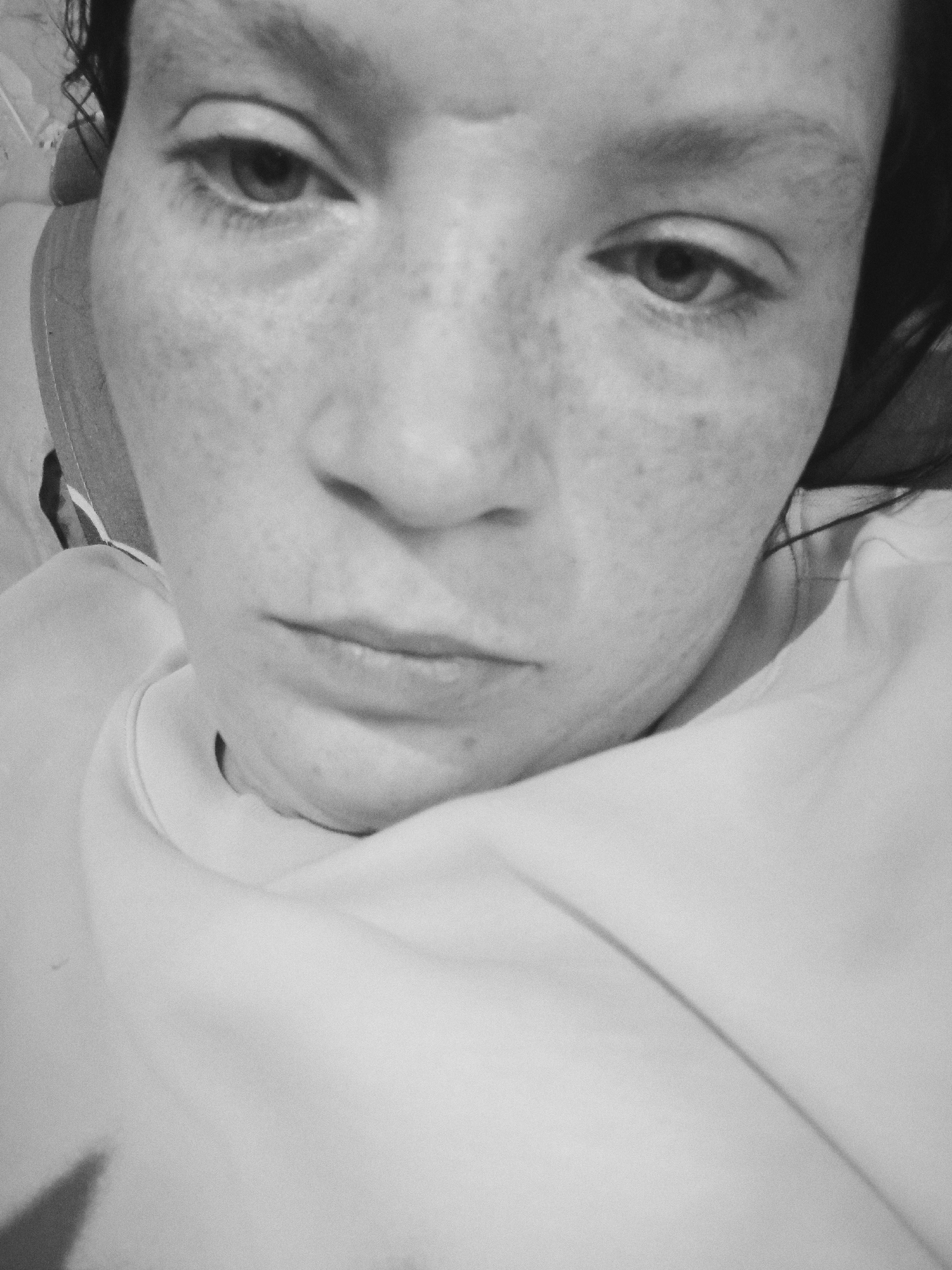 The image is a close-up, black and white photograph of a person's face, taken from a low angle. The subject is looking downwards, with their eyes partially closed. Freckles are visible on the skin. The person is wearing a light-colored garment, possibly a t-shirt or sweatshirt. The background is out of focus and appears to be indoors. There is no identifiable information in the image that would place it in Dalneretschensk, Russia, or any other specific city.
