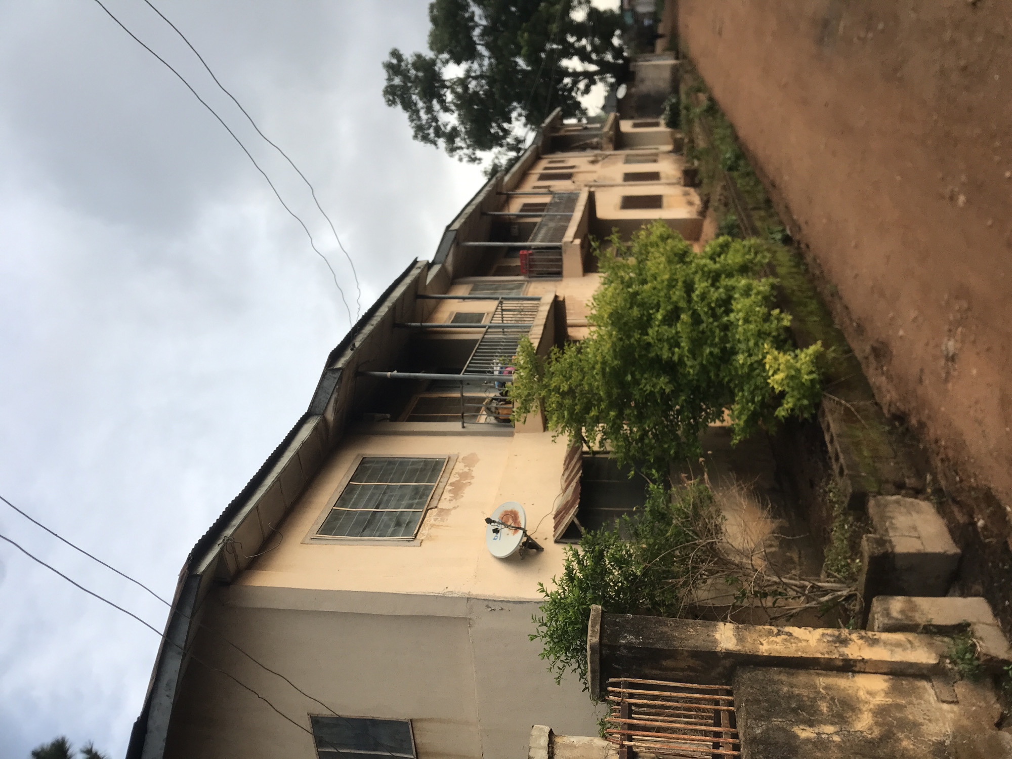 This image depicts a multi-story building and its immediate surroundings in Rafin Par, Nigeria, under a heavily overcast sky. The primary subject is a tall, yellowish-beige building, possibly an apartment complex or a large family house, extending vertically through the frame. It features a corrugated metal roof and multiple floors, each equipped with windows, some secured with metal grilles. Several balconies or verandas with metal railings are visible on the upper levels, with what appears to be laundry hanging on one. A small, weathered white satellite dish is mounted on the side of the building on a lower floor, near a window.
To the right of the building, a reddish-brown dirt road or path stretches into the distance, appearing unpaved and uneven. Green foliage is abundant, with lush bushes lining the edge of the dirt path and a large tree towering in the background behind the building. In the foreground on the right, a low concrete wall or fence, partially covered by vegetation, separates the building's area from the road, with a rusty metal gate visible at its base. Overhead, two thin utility wires are prominent against the uniformly grey sky. The scene is static, devoid of any human activity. The cloudy conditions suggest daytime, likely under a humid or rainy weather pattern. No visible text is discernible in the image.
