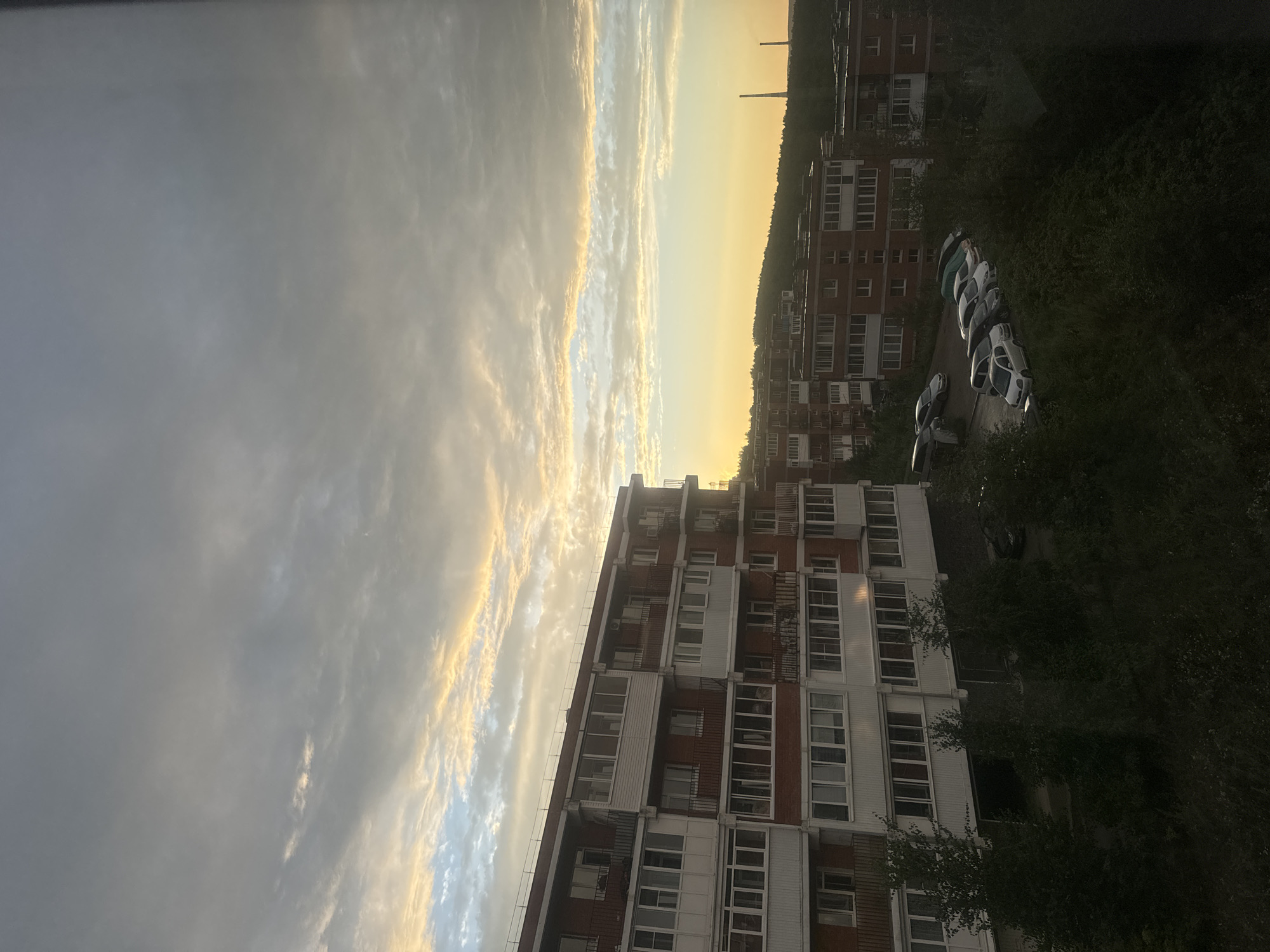 The image shows an overcast sky with a band of golden light near the horizon, indicating either sunrise or sunset. In the foreground, there are several apartment buildings, likely in the city of Markova, Russia. The buildings are multi-story structures with red brick exteriors and white balconies or enclosed sunrooms. Some trees and a parking lot with several cars are visible at the base of the buildings. In the distance, there are more buildings and a hill covered in trees. Two tall, thin industrial chimneys are visible on the right side of the frame, silhouetted against the sky. The overall impression is of a residential urban or suburban area at twilight.