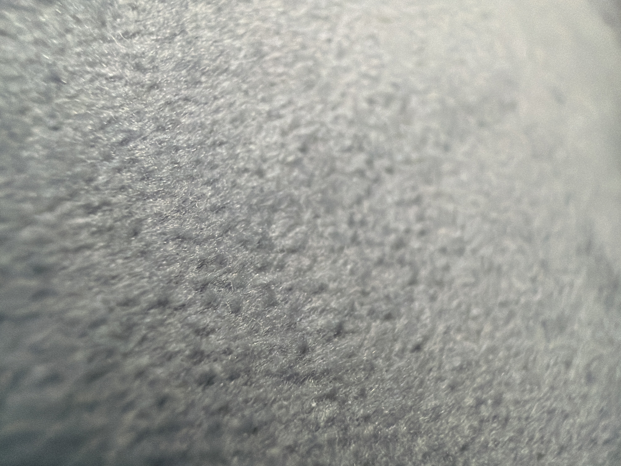 The image shows a close-up, abstract view of a textured surface. The surface appears to be made of fine fibers, creating a fuzzy or plush appearance. The texture is uneven, with slight variations in depth and contour. The color is a muted, cool tone, primarily shades of grey with hints of blue. There are no discernible people, objects, or specific environmental features visible. The scene is devoid of any context that would suggest a location within the city of Elin Pelin, Bulgaria.