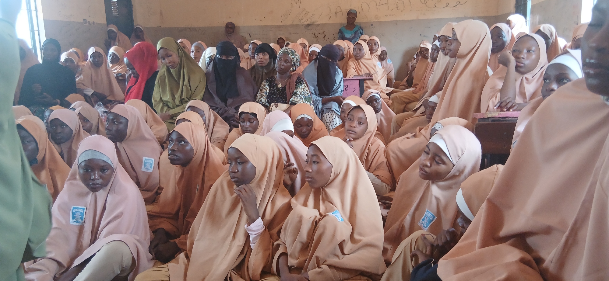 A group of young people, mostly women and girls wearing light brown hijabs and gowns, are seated on the floor in a room. Some are looking forward, while others are looking down. The room appears to be a classroom, with a blackboard on the wall in the background that has some writing on it. The lighting suggests it is daytime, possibly in Nigeria, given the location information provided. The general atmosphere is one of a gathering or a lesson taking place.