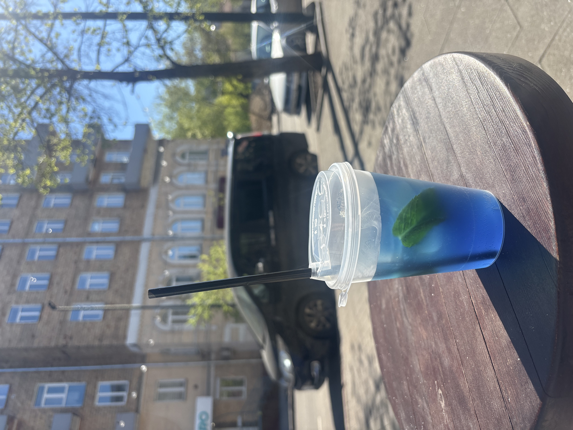 The image captures a blue drink in a clear plastic cup with a lid and black straw, resting on a round, dark-colored wooden table. A green leaf is visible inside the drink. In the background, a multi-story building with numerous windows is partially visible, alongside parked cars and trees with budding leaves, suggesting it is likely spring. The image appears to be taken from a position that shows the street view through a reflective surface, possibly a window. Given the location constraint, this could potentially be in Krasnoyarsk, Russia, however, there's no specific information in the image to directly confirm it.
