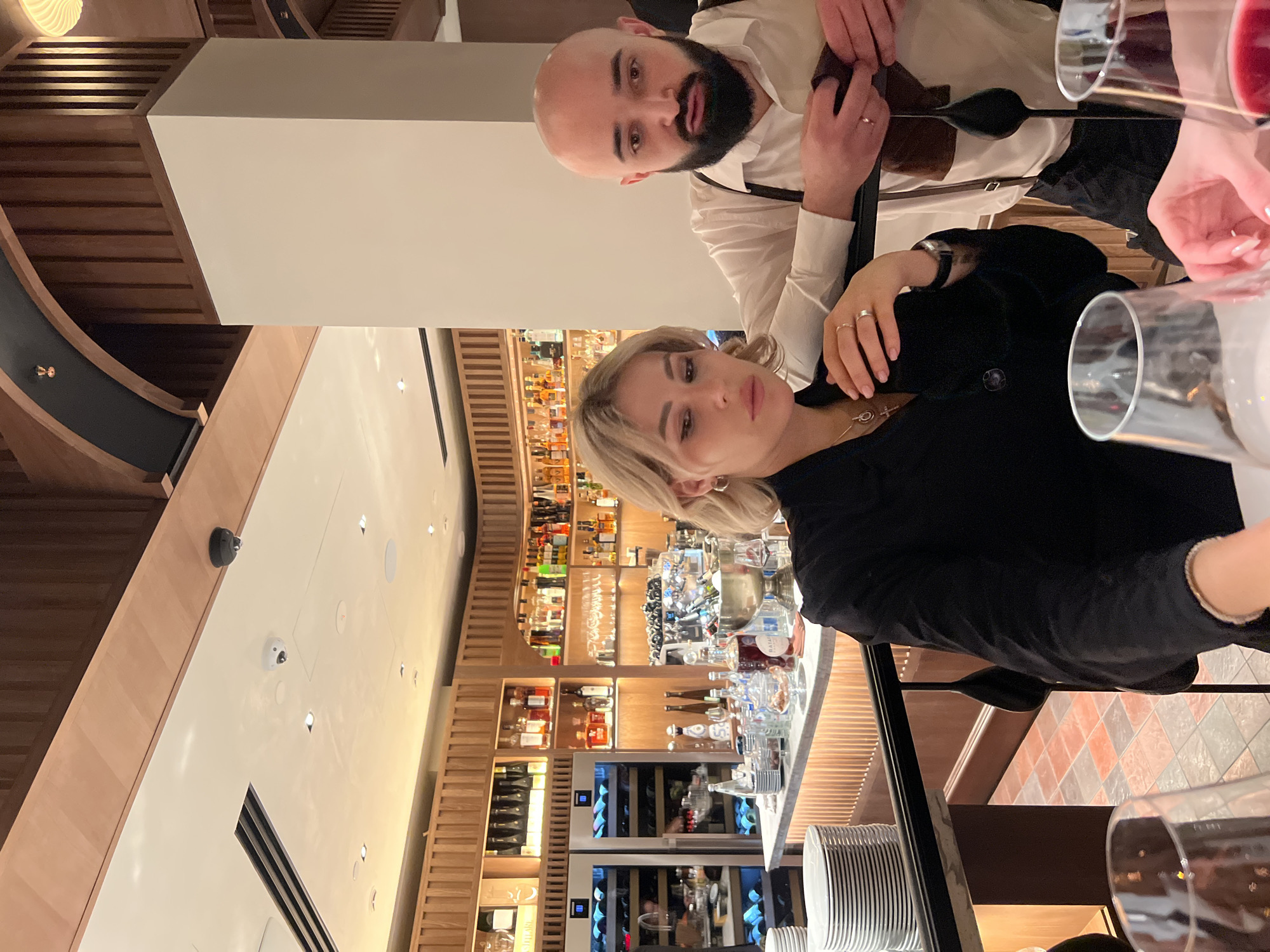 An indoor photograph captures two individuals, a man and a woman, seated at what appears to be a bar or table in an upscale restaurant or bar in Moscow, Russia. The image is taken at eye level, presenting a casual pose.
The man, positioned in the upper portion of the frame (when oriented correctly), is bald with a prominent dark beard and mustache. He wears a white long-sleeved shirt and black suspenders, looking directly at the camera with a neutral expression. A ring is visible on his left ring finger, and his hands are clasped, holding a dark, flat object near his chest.
The woman, in the lower portion, has blonde, shoulder-length hair and is looking slightly off-camera with a serene expression. She wears a black top or jacket and a necklace with a small pendant. Rings adorn her right ring finger and left index finger, and she wears a watch on her left wrist. Her fingernails are painted a light nude color.
The setting features a stylish interior with extensive wooden paneling and slatted architectural elements. Behind the woman is a well-stocked bar with numerous bottles of various spirits and wine displayed on wooden shelves. Integrated wine coolers are also visible. One legible bottle label reads "BATIDA DE COCO". The ceiling features recessed lighting and security cameras. In the background, a curved dark railing suggests multiple levels or a unique architectural design. The flooring visible in parts of the image consists of reddish-brown and gray square tiles. In the foreground, wine glasses, one containing red liquid, are placed on a dark surface, and a stack of white plates is seen on a lower shelf.
The artificial lighting suggests the event is taking place in the evening or at night. The individuals appear to be posing for a photograph during a social outing.