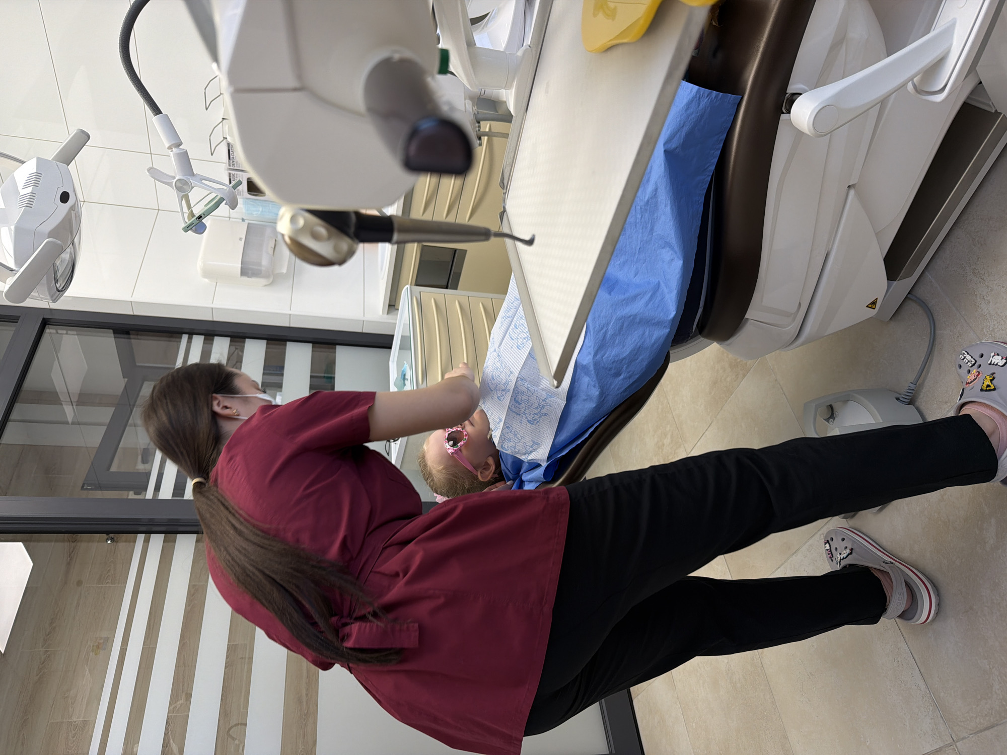 A dental examination is taking place in what appears to be a modern dental clinic. A young child is lying in a dental chair, wearing pink glasses. A dental professional, wearing a maroon scrub top and black pants, stands beside the chair, holding a dental instrument. The room is equipped with standard dental equipment, including a dental light and a tray with tools. The clinic has a clean and well-lit environment. The city of Kamyshin, Russia, cannot be confirmed as the location based on the provided image.