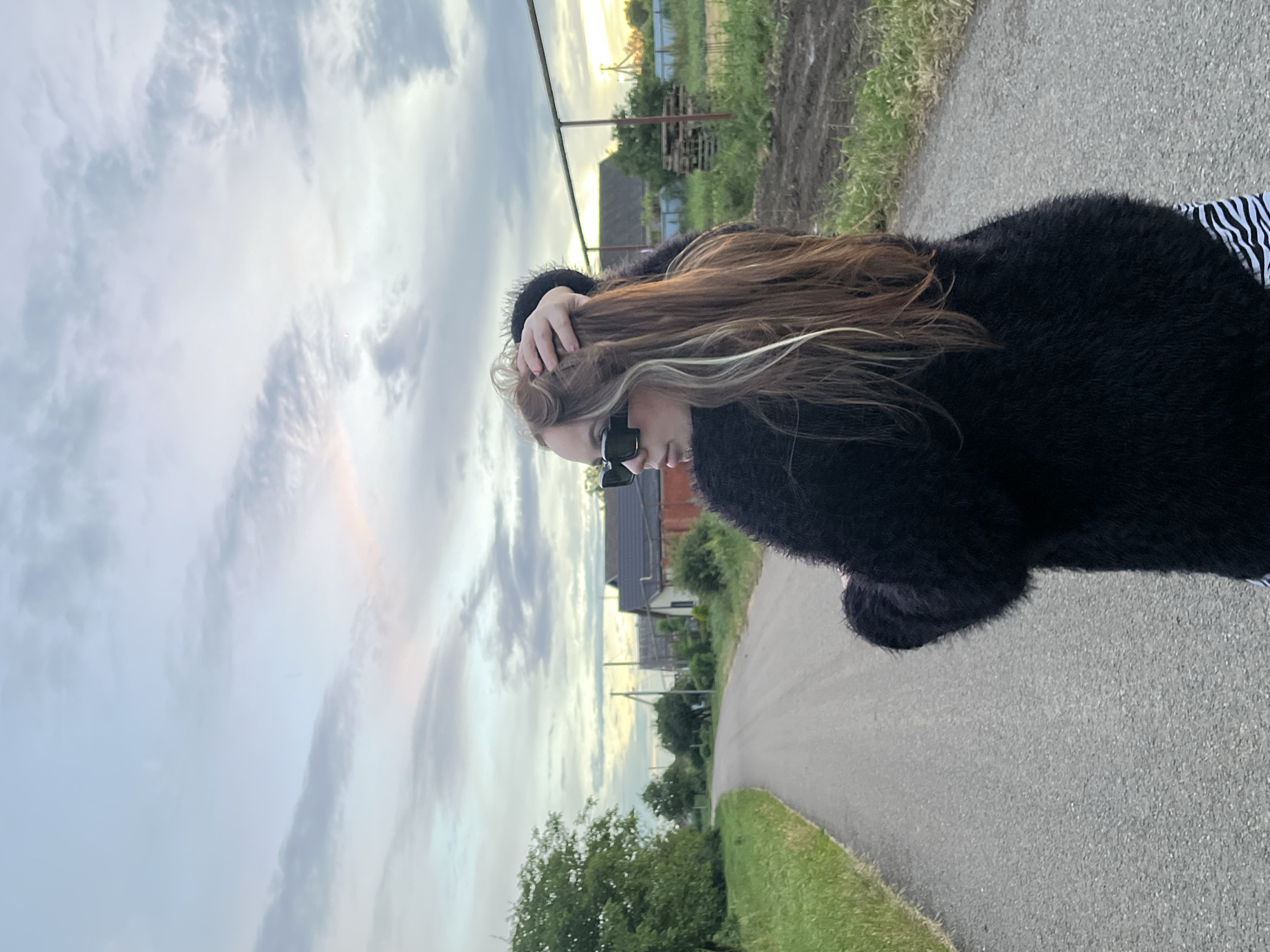 An adult female is standing on an asphalt road in a rural area, possibly in Novopokrovskaya, Russia. She is wearing a black fluffy sweater and sunglasses, with her hand in her hair. The road curves to the left, with grassy fields and trees on the left side. On the right side of the road, there are houses, a fence, and stacks of wooden pallets. The sky is overcast with clouds and a hint of sunset colors. The overall scene suggests a casual, outdoor setting during dusk.