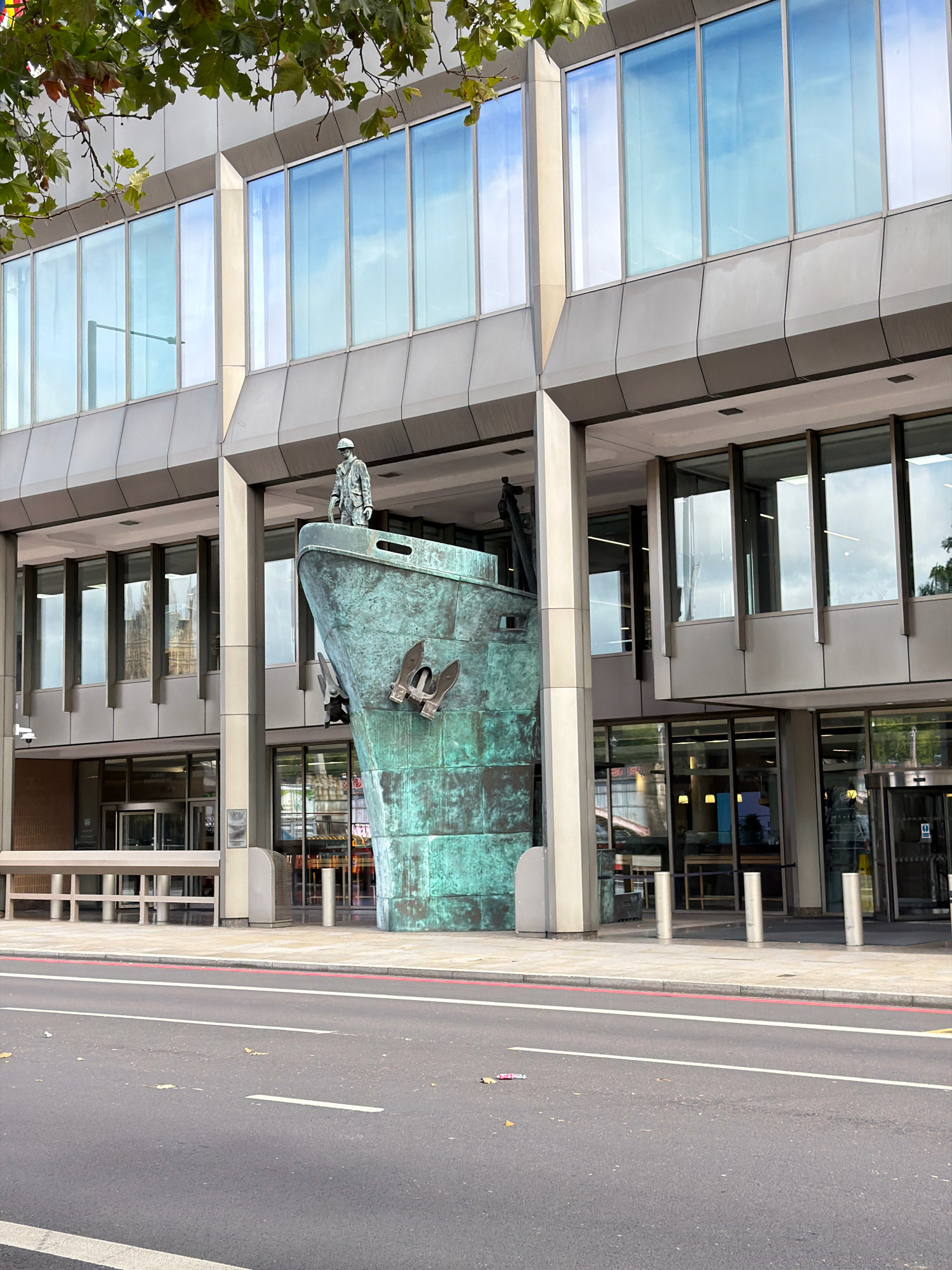 An exterior medium shot captures a large, verdigris-patinated ship's prow sculpture positioned outside a modern building in London, United Kingdom. The sculpture, which appears to be made of bronze, features a figure standing on its deck and two anchors attached to its bow. The building behind the sculpture is a multi-story structure with a facade of reflective windows and grey architectural panels. The street in front of the building is paved with asphalt and has white and red painted lines indicating lanes and a sidewalk. The overall lighting suggests it is daytime with overcast skies, as evidenced by the soft, diffused light and the reflections on the windows. The scene is static with no people or vehicles actively engaged in movement.