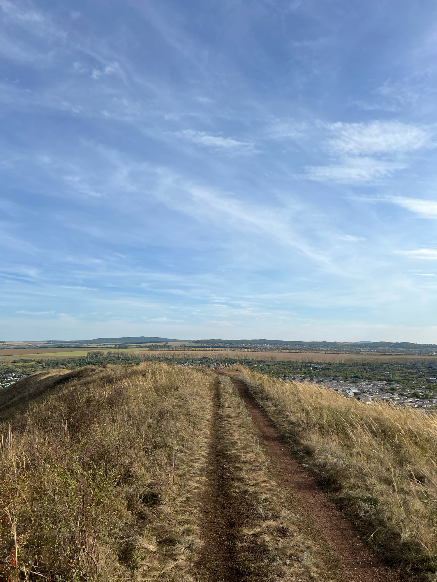 The image depicts a view from a grassy hilltop overlooking a city. A dirt path winds its way up the hill, covered in dry grass and sparse vegetation. In the distance, a city with buildings and roads is visible, nestled between rolling hills. The sky above is a clear blue, with wispy clouds scattered across it. The overall impression is one of a natural landscape meeting a populated area under a bright sky. The location is the city of Ishimbay, Russia, a city known for its oil production and situated in the Republic of Bashkortostan. The scene captures a serene, natural vista, with the city providing a backdrop to the undeveloped hilltop.