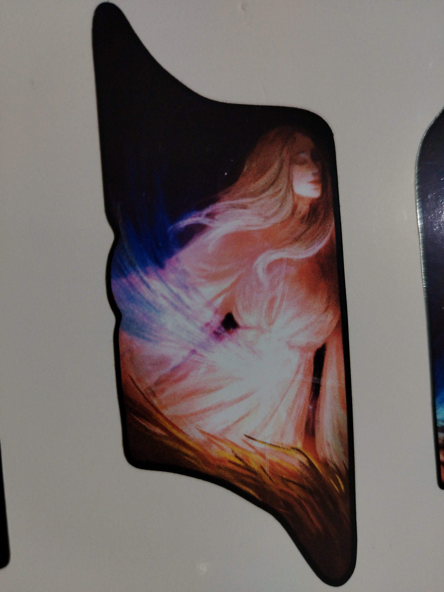 The image is a close-up of a die-cut sticker or magnet featuring an ethereal figure. The figure has long, flowing blond hair and is draped in what appears to be a sheer or glowing garment. The background is dark, possibly suggesting a night sky with blue and purple hues near the figure. The figure is depicted in a soft, painterly style with warm tones around the lower portion. The sticker or magnet has a dark, rounded edge. There is no indication of the location being Izhevsk, Russia, or any other city.