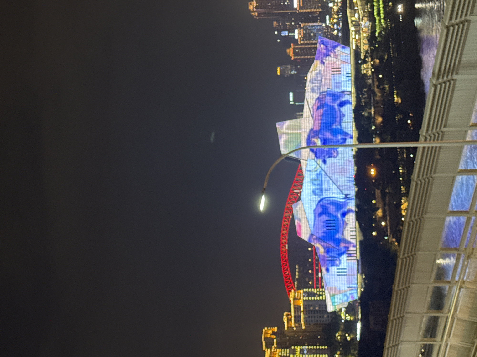 This is a nighttime photograph of a modern architectural building in Yuzhong District, China. The building's facade is illuminated with a light show, prominently featuring a large, stylized blue bull. Behind the building, a cityscape of illuminated skyscrapers is visible. In the foreground, a bridge railing with reflective surfaces leads the eye toward the illuminated structures. A streetlamp is positioned to the right of the frame. The sky is dark, indicating nighttime.