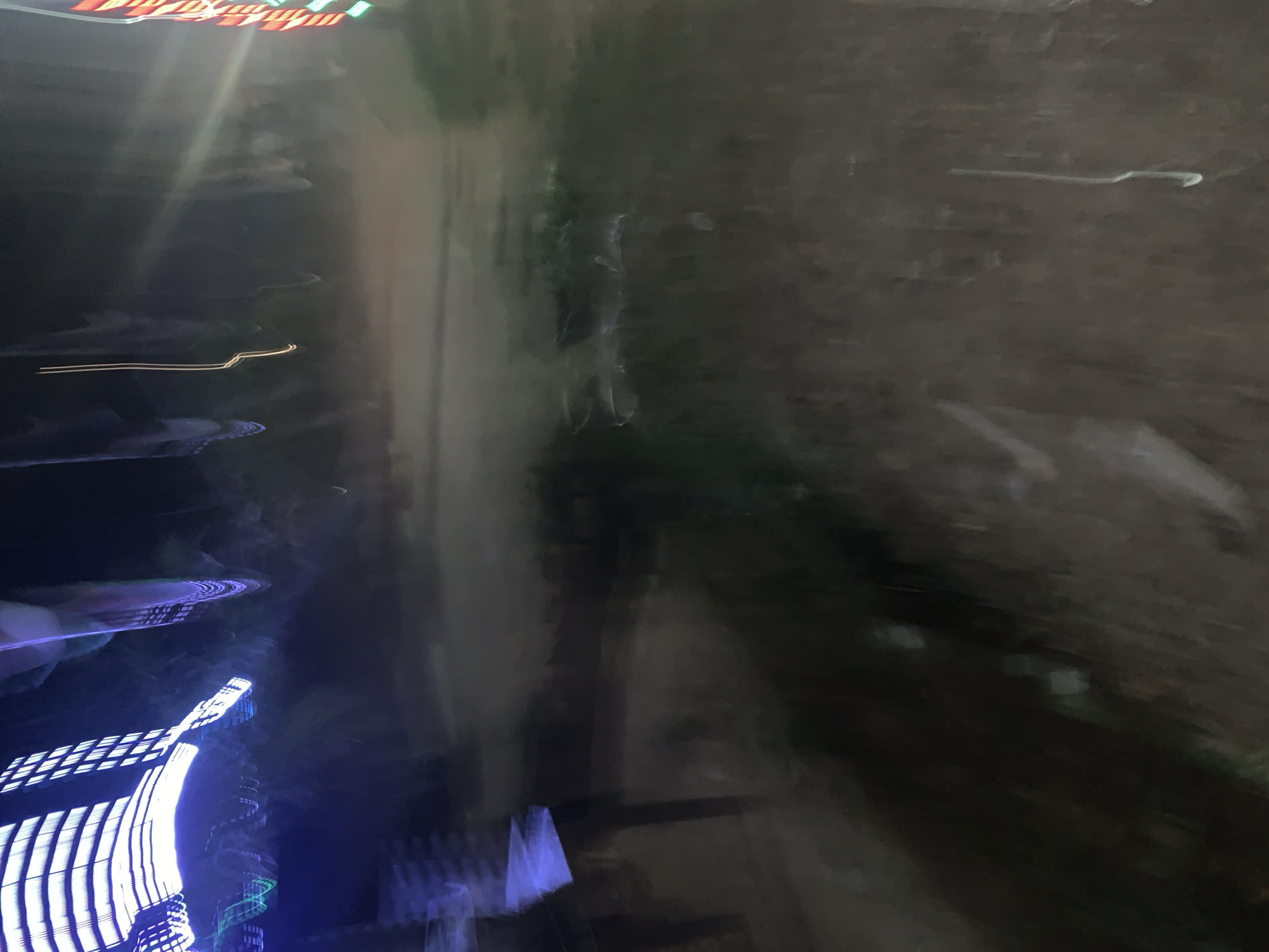 This is a photograph taken at night, likely outdoors, as suggested by the presence of what appears to be a path or unpaved ground on the right side of the frame. The image is abstract and heavily affected by motion blur and light trails, making it difficult to discern specific details.
On the left side of the image, there are bright, colorful light trails that suggest artificial lighting, possibly from vehicles, signs, or fireworks. Some of these trails are streaks of red, green, and white. Further down, there are also blue and white light streaks. It's possible these lights are from a fairground or a busy urban area at night, or perhaps a moving vehicle with its lights on.
The central and right portions of the image are darker and more muted, with indistinct shapes and textures. There are some faint, wispy light streaks or reflections visible in this area as well. The overall impression is one of movement and a somewhat chaotic or energetic atmosphere due to the light streaks. The time of day is clearly night due to the darkness and the nature of the light sources. No specific weather conditions can be determined. No people or specific objects are clearly identifiable, and there is no visible text. The context provided places the scene in Gura Top, Nigeria.