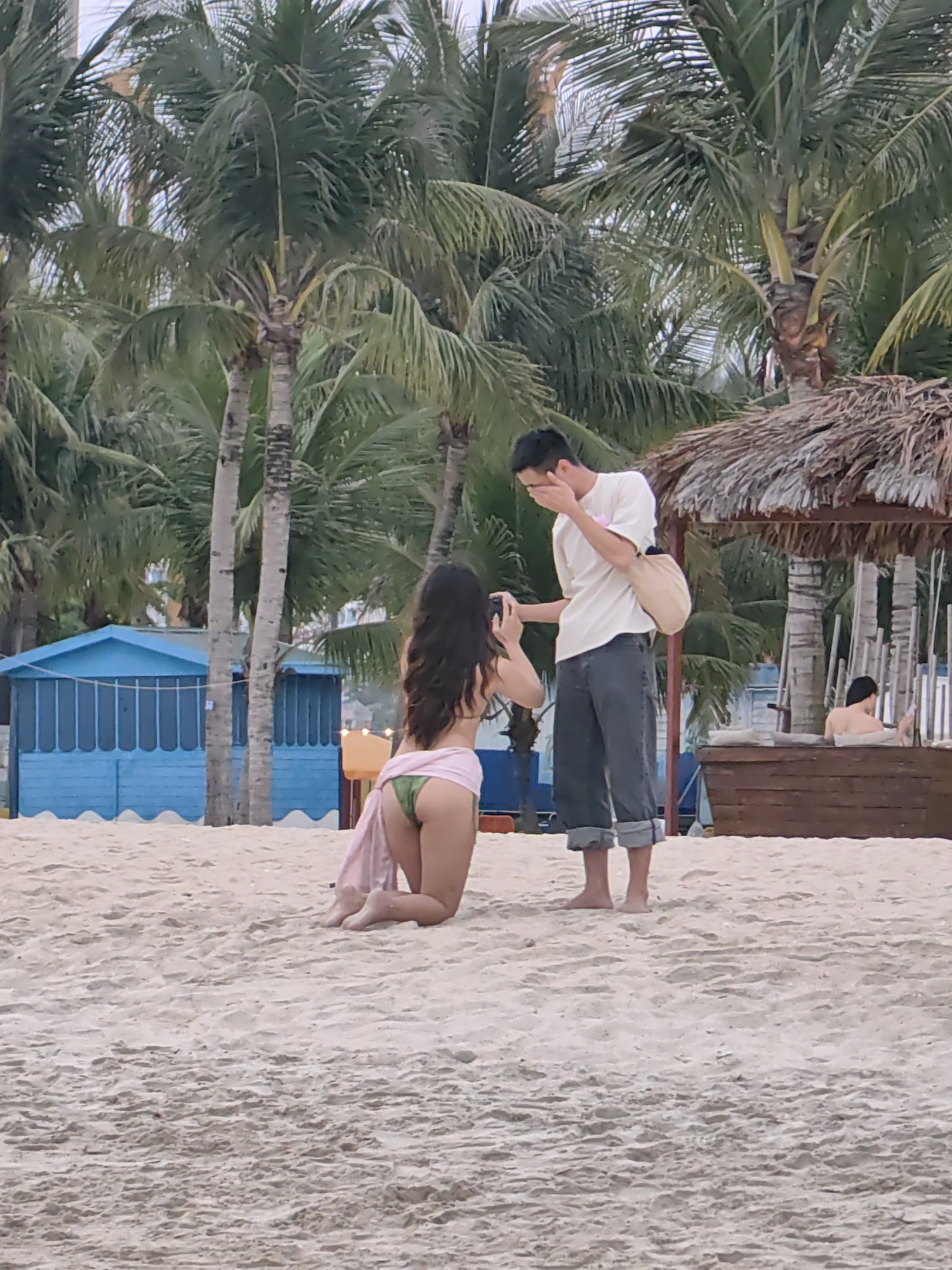The image captures an intimate moment on a sandy beach, likely in Bai Chay Ward, Vietnam. In the foreground, a woman is kneeling on the light-colored sand, facing a man. She has long dark hair, wears a green bikini bottom, and a pink sarong or wrap draped around her waist. She holds a black camera, seemingly taking a picture or filming the man.
The man stands barefoot before her, dressed in a light-colored t-shirt, dark rolled-up pants, and a light-toned shoulder bag. He covers his face with one hand, indicating a strong emotional reaction, possibly surprise, joy, or overwhelmed feeling.
The background is dominated by numerous tall palm trees, reinforcing the tropical beach setting. To the left, a small blue wooden hut with white trim is visible. On the right, there's a thatched-roof structure, possibly a sunshade, and a wooden boat-shaped seating area where another person is partially visible. The soft, diffused lighting suggests the time of day is either early morning or late afternoon. No visible text is present in the image.