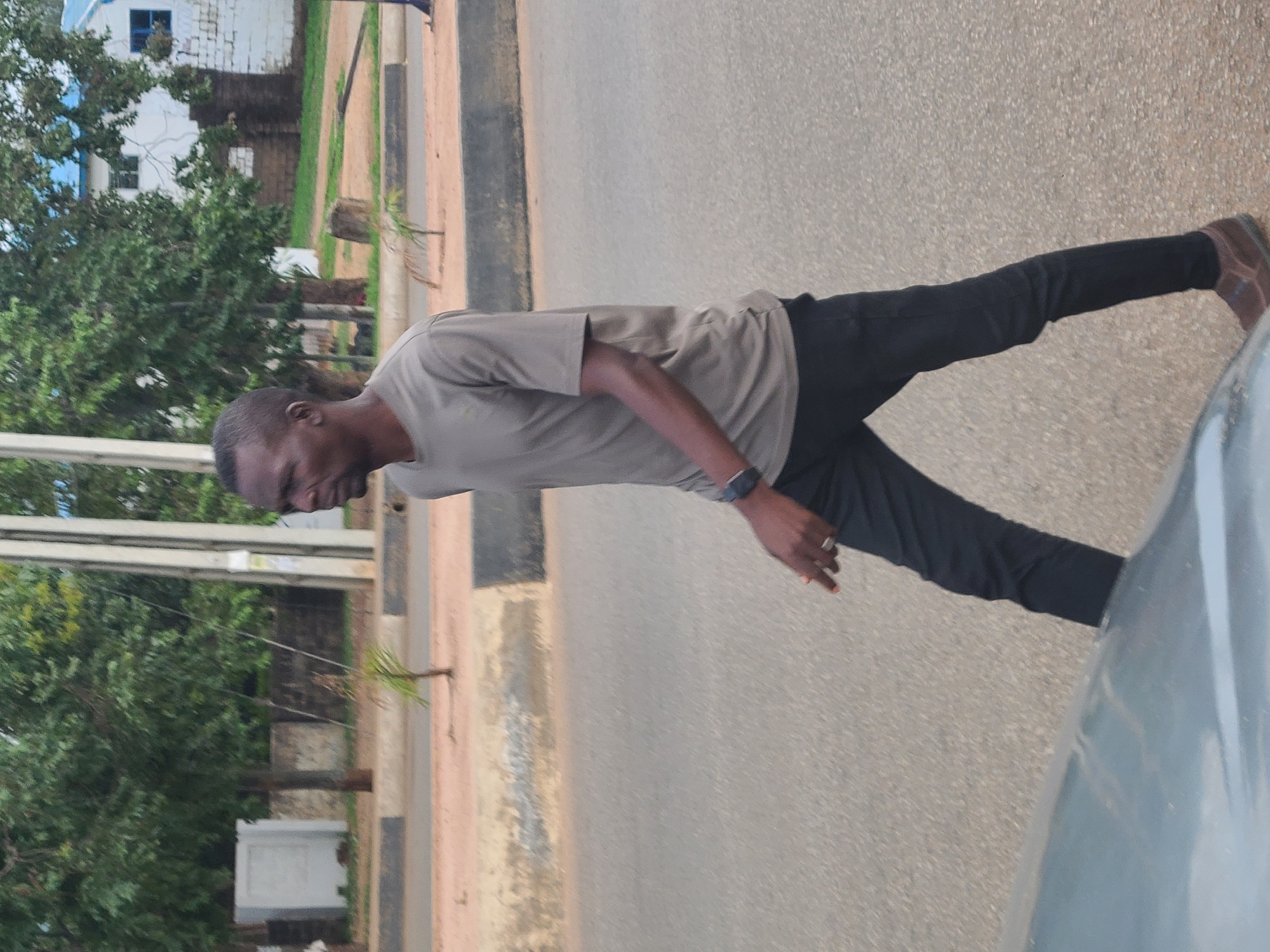 A man in a light-gray t-shirt, black pants, and brown shoes stands on an asphalt road. He is also wearing a watch. To his left is a sidewalk and a grassy area with trees. On the left of the grassy area is a building. To his right is a car. The location appears to be an urban setting.