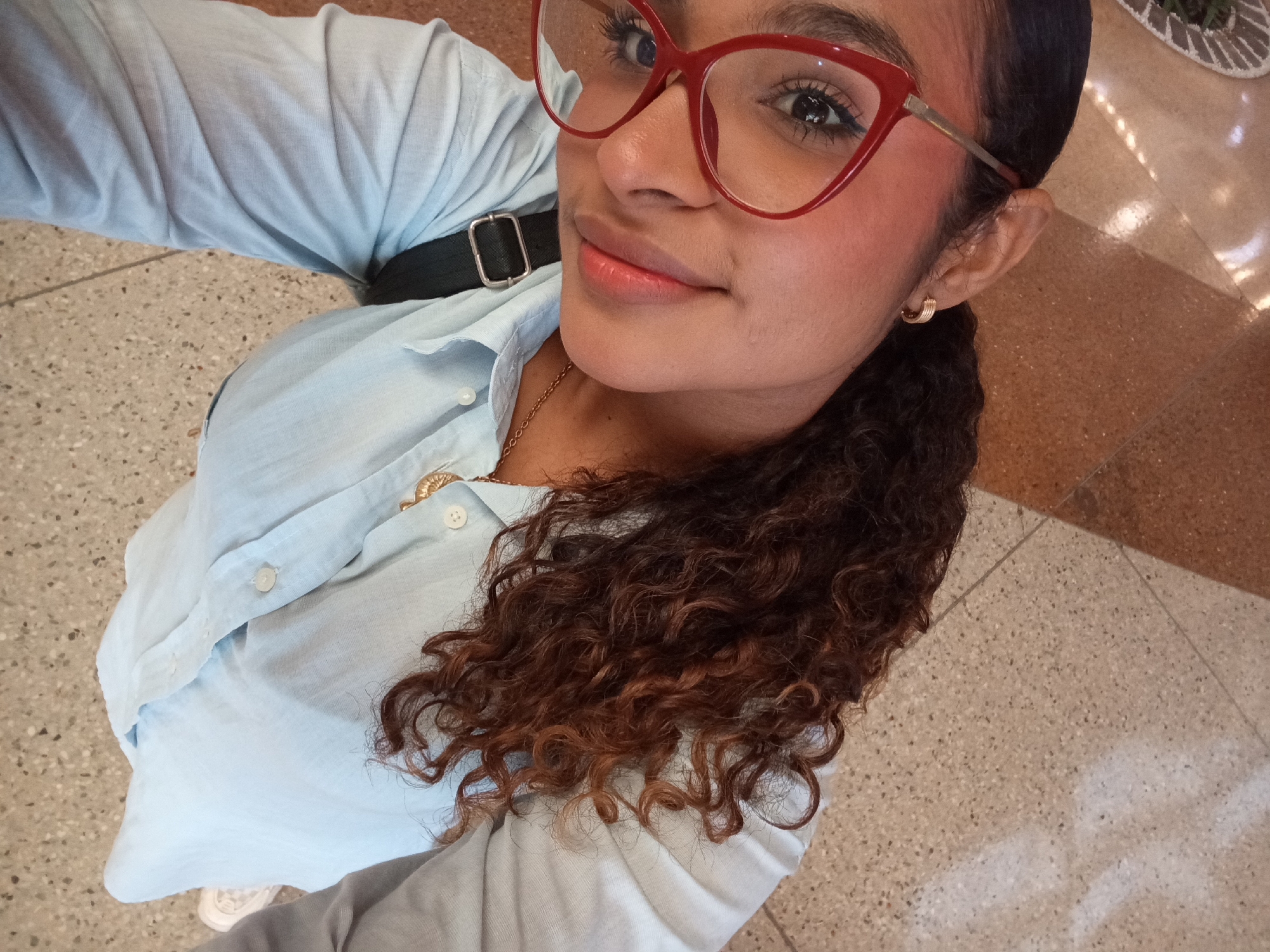 A high-angle selfie captures a woman looking directly at the camera with a pleasant, subtle smile. She has a medium skin tone and dark, curly hair with visible reddish-brown highlights, cascading over her right shoulder. Her eyes are framed by bold red cat-eye glasses and accentuated with eyeliner. She wears subtle makeup, including blush and a pink lip tint. Gold hoop earrings adorn her ears, and a delicate gold chain necklace with a circular pendant is visible around her neck.
She is dressed in a light blue button-down shirt, and a dark, possibly black, strap—likely from a bag—crosses her left shoulder. The scene is indoors, characterized by a speckled, terrazzo-like tiled floor in shades of beige and brown, featuring some lighter patterns or reflections. The lighting suggests it is daytime. The woman appears calm and relaxed. No visible text is present in the image. The location context is Pampatar, Venezuela.