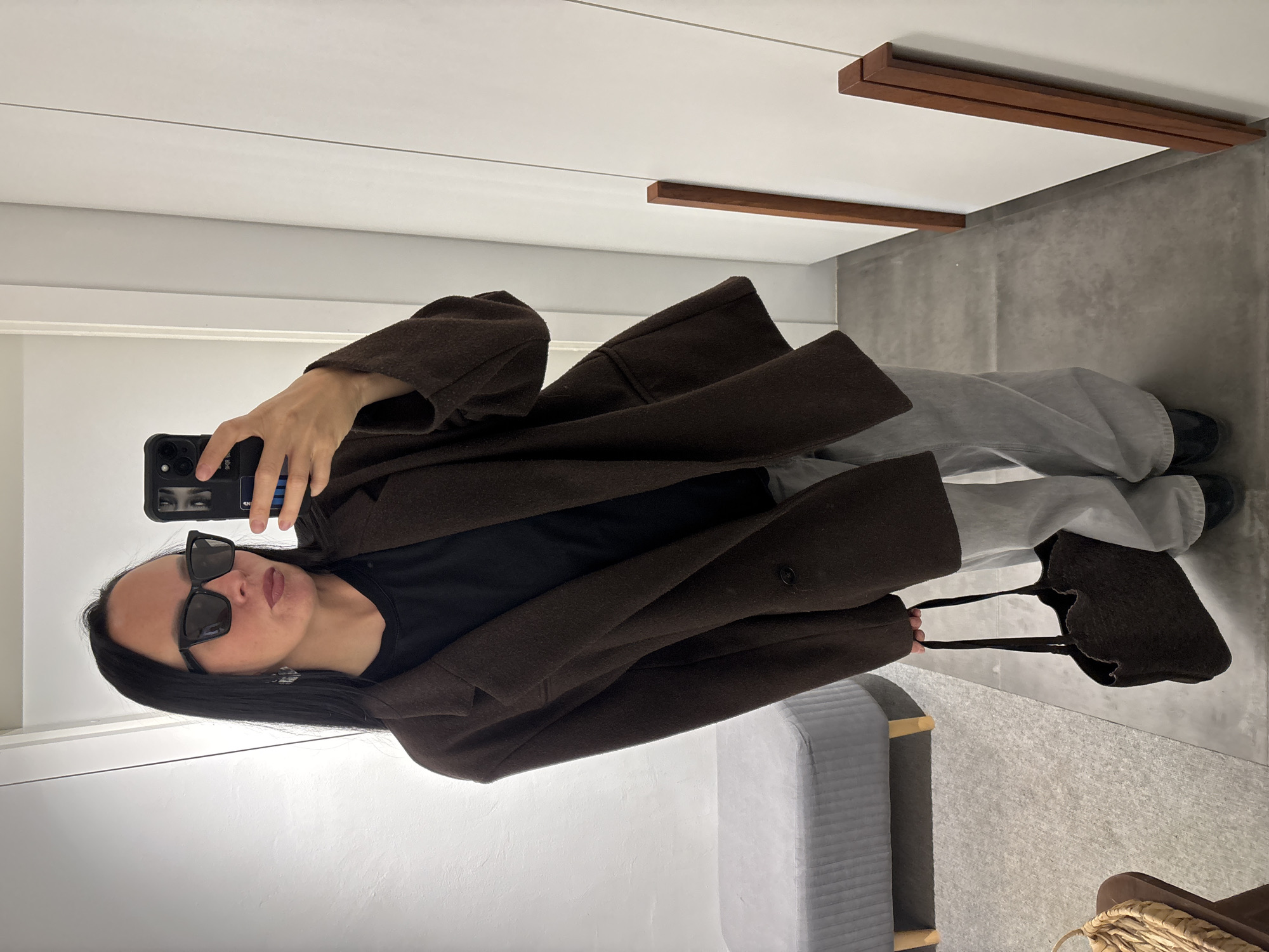 A female subject is posing for a selfie in what appears to be an interior space. She is wearing sunglasses, a dark T-shirt, gray sweatpants, and a brown coat, and she is holding a brown tote bag. The background includes white walls, gray floors, and some furniture including a gray bench and a wooden shelf-like structure. The location is unknown.