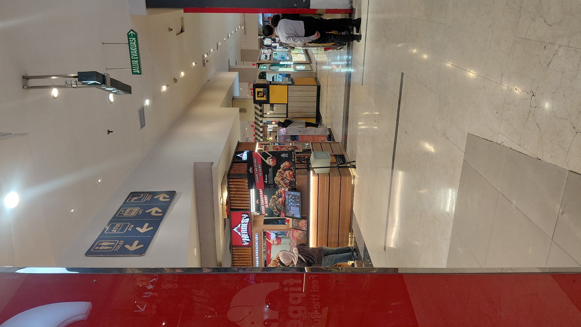 The image depicts a well-lit interior, likely a shopping mall or a similar commercial establishment, in Purwokerto, Indonesia. The floor is tiled with a marble-like pattern, reflecting the overhead lighting.
On the left side of the frame, a portion of a large red structure is visible, possibly part of a store facade. Further into the scene, a food stall is set up, featuring a prominent sign with the logo "HEROES" and advertisements for "OISHI TAKOYAKI". The stall has a wooden counter and display screens showing menu items. A person wearing a hijab and casual clothing is standing at the counter, appearing to interact with the stall.
In the background, several other commercial units are visible, along with more patrons. A sign indicating "JALUR EVAKUASI" (Evacuation Route) with an arrow pointing right is mounted on the ceiling, suggesting safety protocols are in place. Another directional sign on the wall displays icons and text for "TOILET," "ATM," and "LIFT," all with directional arrows. Two individuals, one in a light-colored collared shirt and dark pants, and another in a darker uniform, stand near a storefront in the mid-ground, facing away from the camera. The overall environment suggests a busy commercial area.
