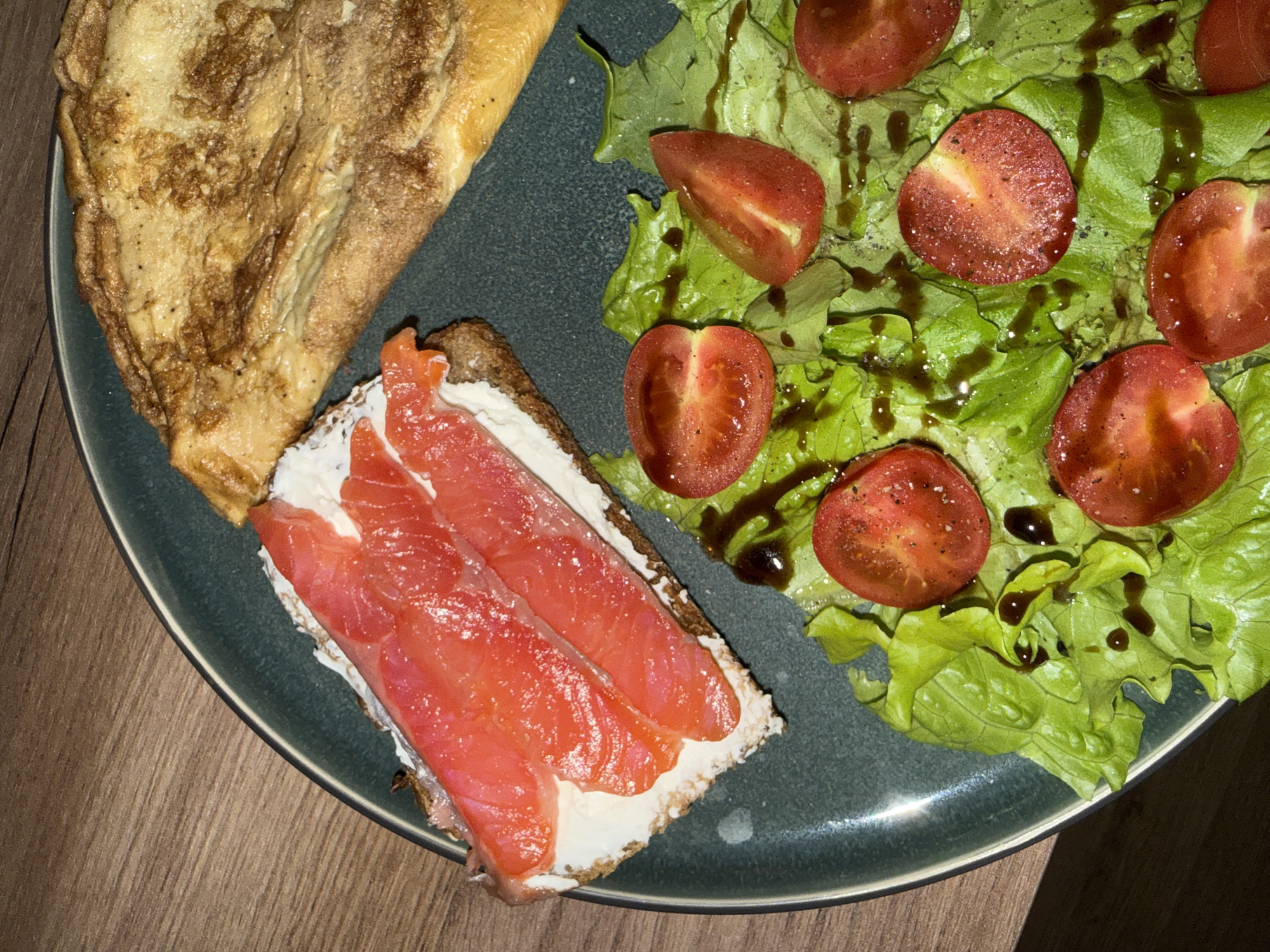 This image presents an overhead view of a well-plated meal, likely a breakfast or brunch, served on a dark grey-blue ceramic plate set on a light brown wooden table. The scene is indoors, in what appears to be a home or restaurant in Krasnoyarsk, Russia.
The plate features three main components. On the upper left, a large, folded item resembling an omelette or thin crepe takes up a significant portion, displaying a golden-brown color with darker, caramelized patches. Below it, towards the center, is a slice of dark bread, possibly rye or whole-grain, generously spread with a thick layer of white cream cheese. This is topped with several thin, vibrant orange-pink slices of smoked salmon, neatly arranged.
The right side of the plate is filled with a fresh, green salad. It comprises crisp lettuce leaves adorned with numerous halved red cherry tomatoes. A dark, glossy dressing, likely balsamic glaze, is artfully drizzled over the salad, and a light scattering of seasoning, possibly salt and pepper, is visible on the tomatoes and greens, adding texture and detail.
The lighting is consistent and bright, illuminating the various textures and fresh colors of the ingredients. No people are visible, nor is any text present in the image. The meal is presented with care, suggesting an inviting and prepared dish ready for consumption.