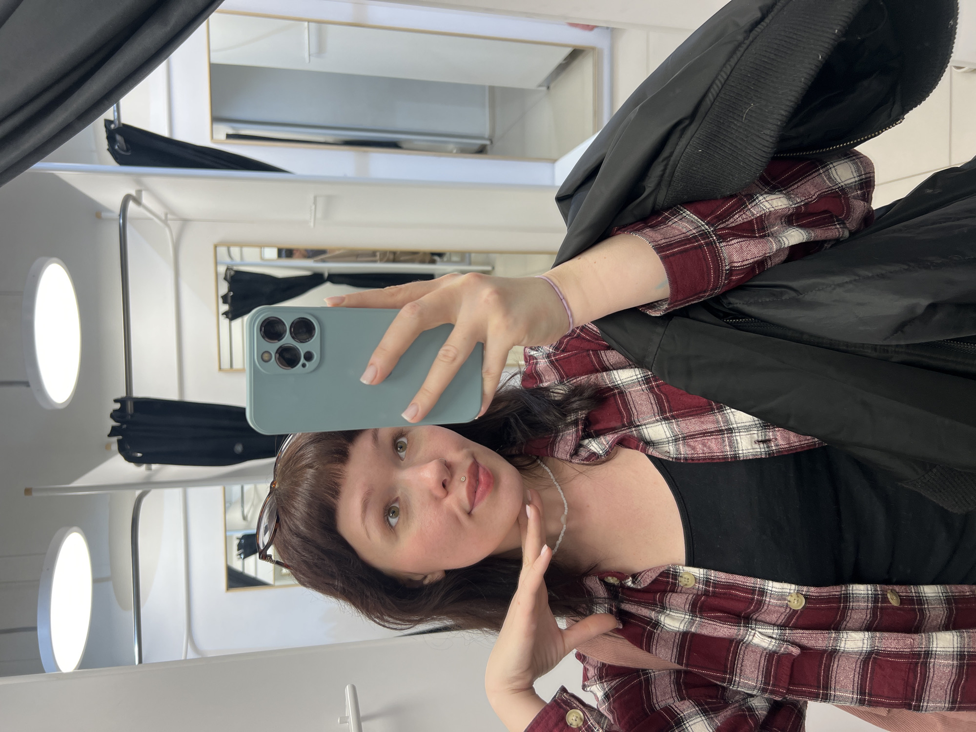 A young woman is captured in a mirror selfie, taken in what appears to be a fitting room in Izhevsk, Russia. The image is oriented with the subject's head tilted to the right, as if the original portrait photo was rotated clockwise.
The woman has dark brown hair with bangs, light eyes, and a lip piercing. She wears a red and white plaid flannel shirt over a black top, with a black jacket draped over her left shoulder. A thin, light-colored bracelet adorns her right wrist, and she has a white beaded necklace. Her fingernails feature a French manicure. She holds a light blue smartphone with her right hand, directing it towards her reflection, and poses with a slight pout and her left hand near her chin, conveying a playful or confident mood.
The setting is characterized by white walls, multiple mirrors with thin frames, and metal hanging rods, consistent with a changing room or retail environment. A dark curtain is visible on the left side of the frame, further suggesting a fitting room. The lighting is artificial, with bright circular light fixtures reflected in the mirrors. No discernible text is visible in the image.