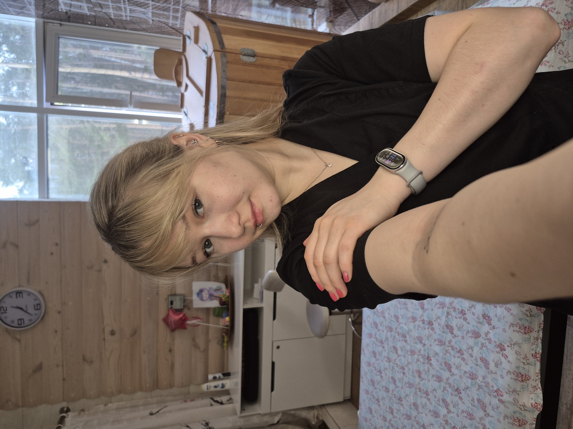 This media file is a rotated selfie of a young woman, likely taken indoors in a rustic setting. The image is rotated 90 degrees counter-clockwise, with the woman's head on the left side of the frame.
**Subjects:** The primary subject is a young woman with blonde hair, fair skin, and light-colored eyes, looking directly at the camera with a neutral to slightly smiling expression. She has multiple piercings in her left ear and a small silver hoop nose ring. She is wearing a black short-sleeved top and a simple pendant necklace. Her fingernails are painted a bright pink/coral color. A grey smartwatch or fitness tracker is visible on her left wrist. A faint tattoo is visible on her upper right arm.
**Objects:** The smartwatch displays "12:36" and the date "10-23 2023". In the background, wooden paneling covers the walls. A window in the upper left shows a blurry view of outdoor trees and bright daylight. A white analog clock, partially visible on the wooden wall in the lower left, shows the numbers 3, 6, 9, and 12, with hands indicating approximately 12:45-1:00. A white storage unit or cabinet is visible in the lower center, and nearby, a small red star-shaped object (possibly a balloon) and a colorful drawing are attached to the wall. A patterned tablecloth or fabric, featuring a light background with pink and blue floral designs, covers a surface in the lower right. A wooden chest or cabinet is partially visible in the upper right.
**Scene/Setting:** The setting is an indoor room with prominent wooden wall paneling, suggesting a cozy, rustic atmosphere. The view through the window indicates a natural outdoor environment, likely with trees.
**Activity or event taking place:** The woman is taking a selfie, holding the camera herself, indicated by her arm's position and direct gaze into the lens.
**Notable details:** The time shown on the smartwatch is 12:36 PM on October 23, 2023. The bright daylight seen through the window confirms it is midday. The overall decor, with wooden walls and rustic furniture, points to a comfortable, possibly rural or vacation home environment. The location context suggests this room is in Chaikovsky, Russia.