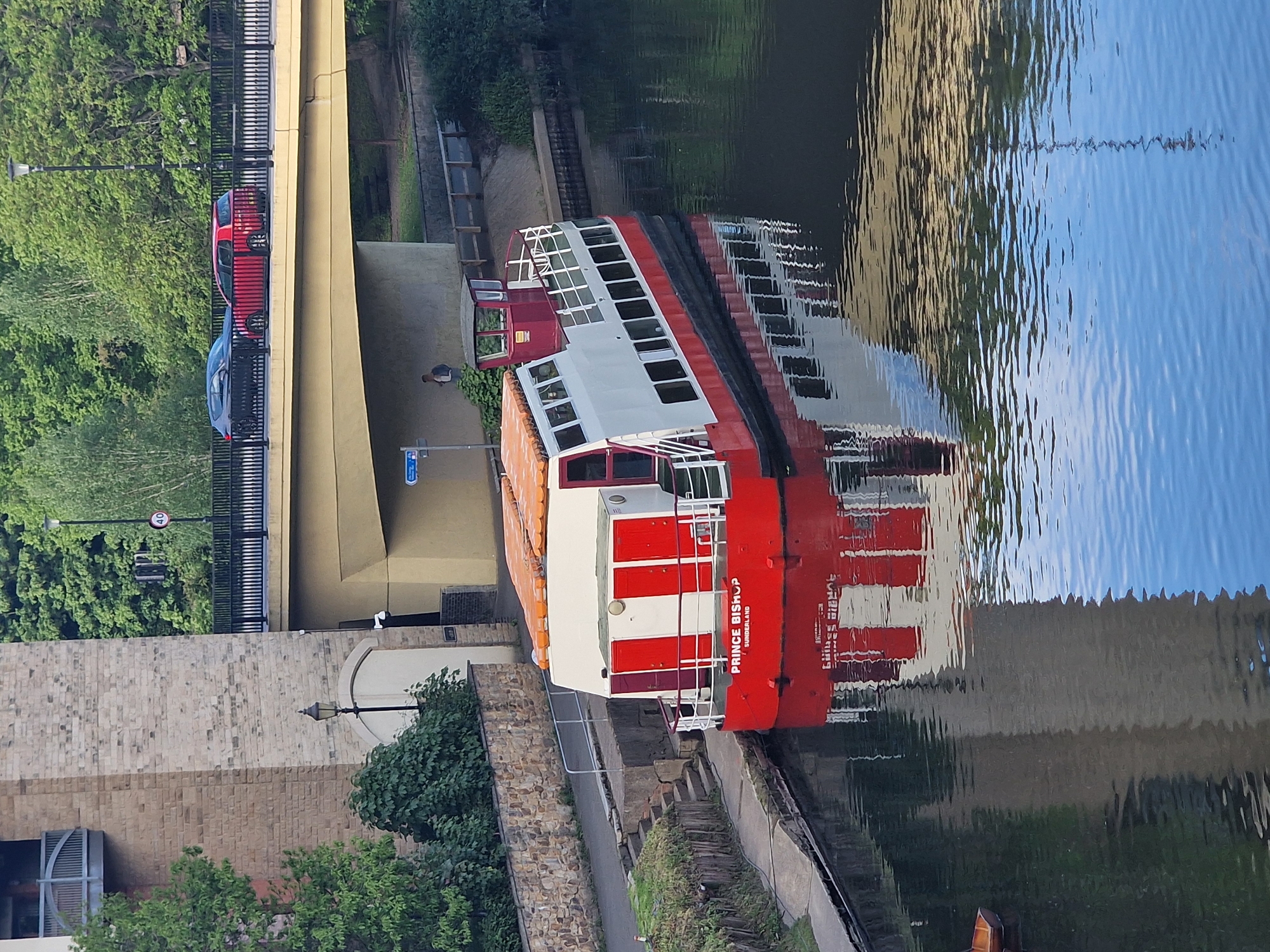 A boat, named "Prince Bishop Sunderland", is docked on a river in Durham, United Kingdom. The boat is white and red and has a glass enclosed top deck. It is docked against a stone and cement embankment. A sidewalk runs along the river bank. Above the boat is a bridge, with two cars driving on it. Trees and bushes are seen along the bank. The river water reflects the bridge and surrounding buildings.