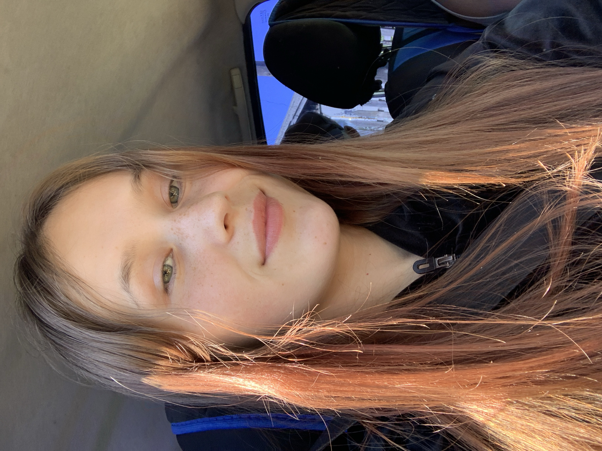 A young woman is seated in a car, her head tilted slightly towards the camera. She has light brown hair with reddish highlights and fair skin with freckles. She is wearing a dark jacket with a visible zipper. The background shows the interior of the car, including a headrest and a glimpse of the outside through the window, revealing a bright sky. The location is not discernible from the image, so it is not possible to confirm if the image was taken in the city of Фоки, Russia.