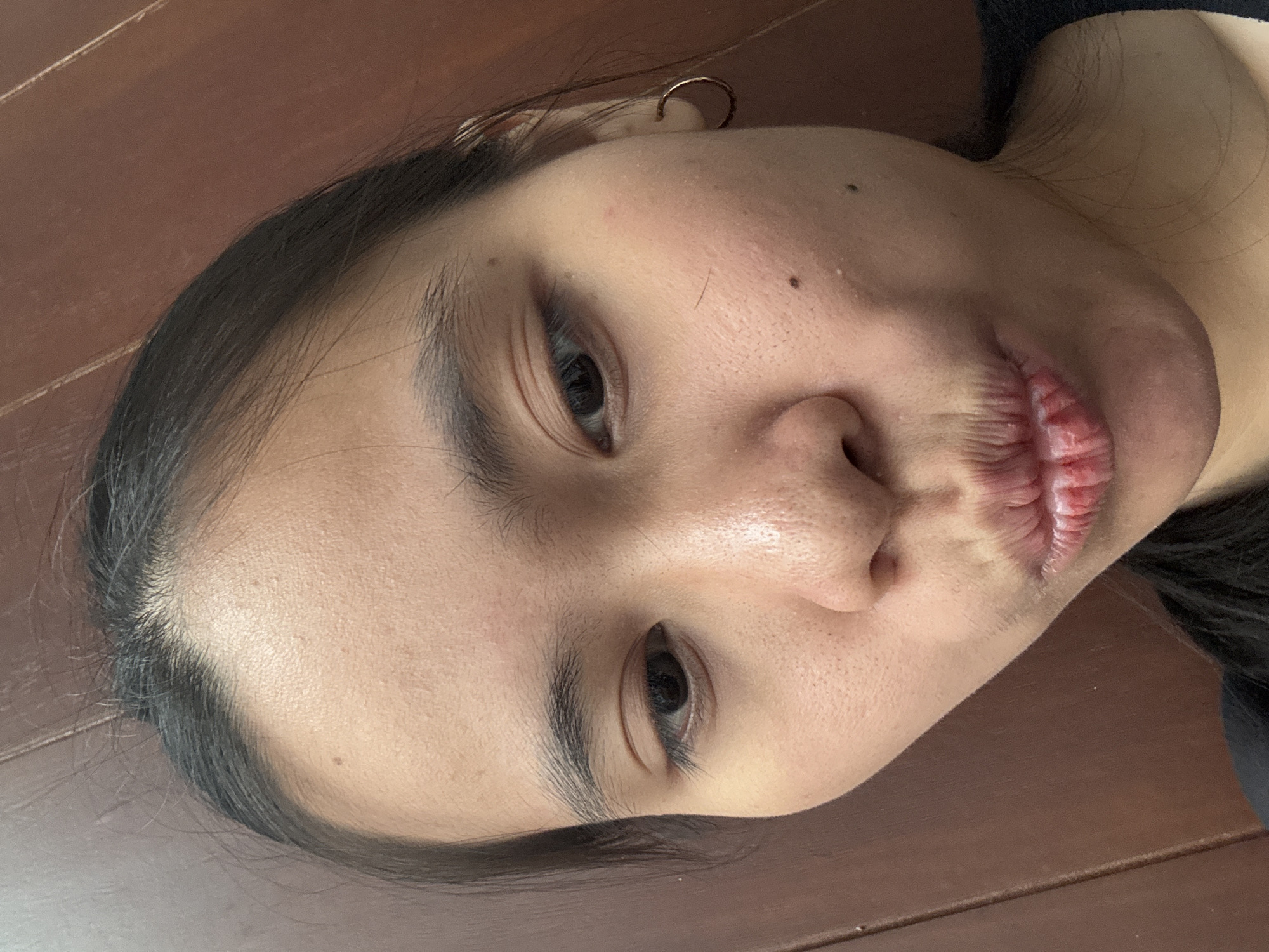 The image is a close-up, selfie-style photograph of a young woman's face. She is looking directly at the camera with her lips pursed in a playful manner. Her skin appears to have visible pores and some blemishes. She is wearing a simple gold hoop earring in her right ear. Her dark hair is pulled back, and some strands frame her face. The background consists of a brown wooden surface, possibly a wall or furniture, with visible wood grain. The lighting appears to be natural and soft, illuminating her face from the front. The image is candid and appears to have been taken indoors. The location is not specified in the image itself.