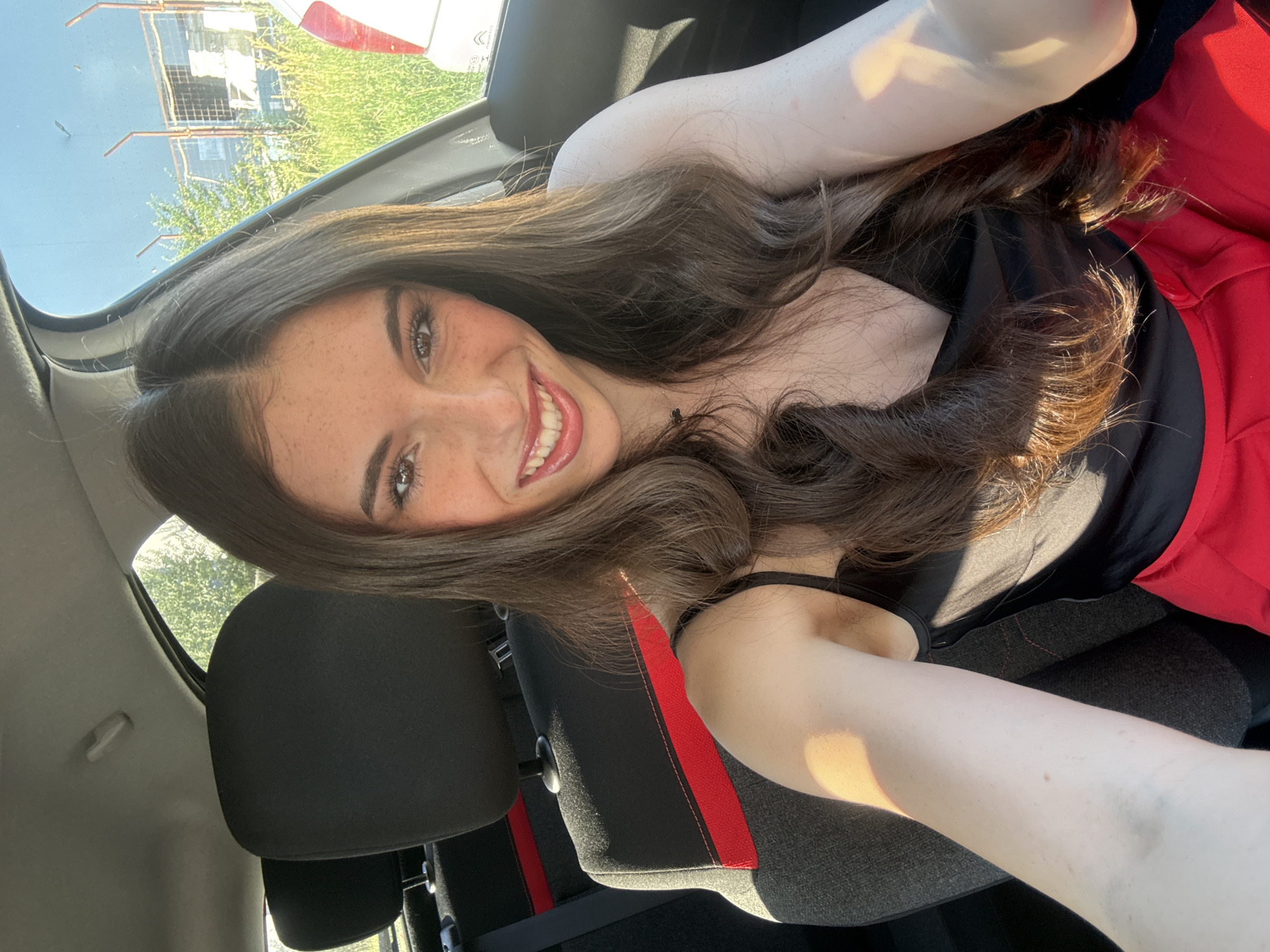 A woman is taking a selfie inside a car. She is wearing a black top and red pants. The interior of the car is black with red accents. The background shows a building and trees through the car window. It is daytime. The location is Plovdiv, Bulgaria.