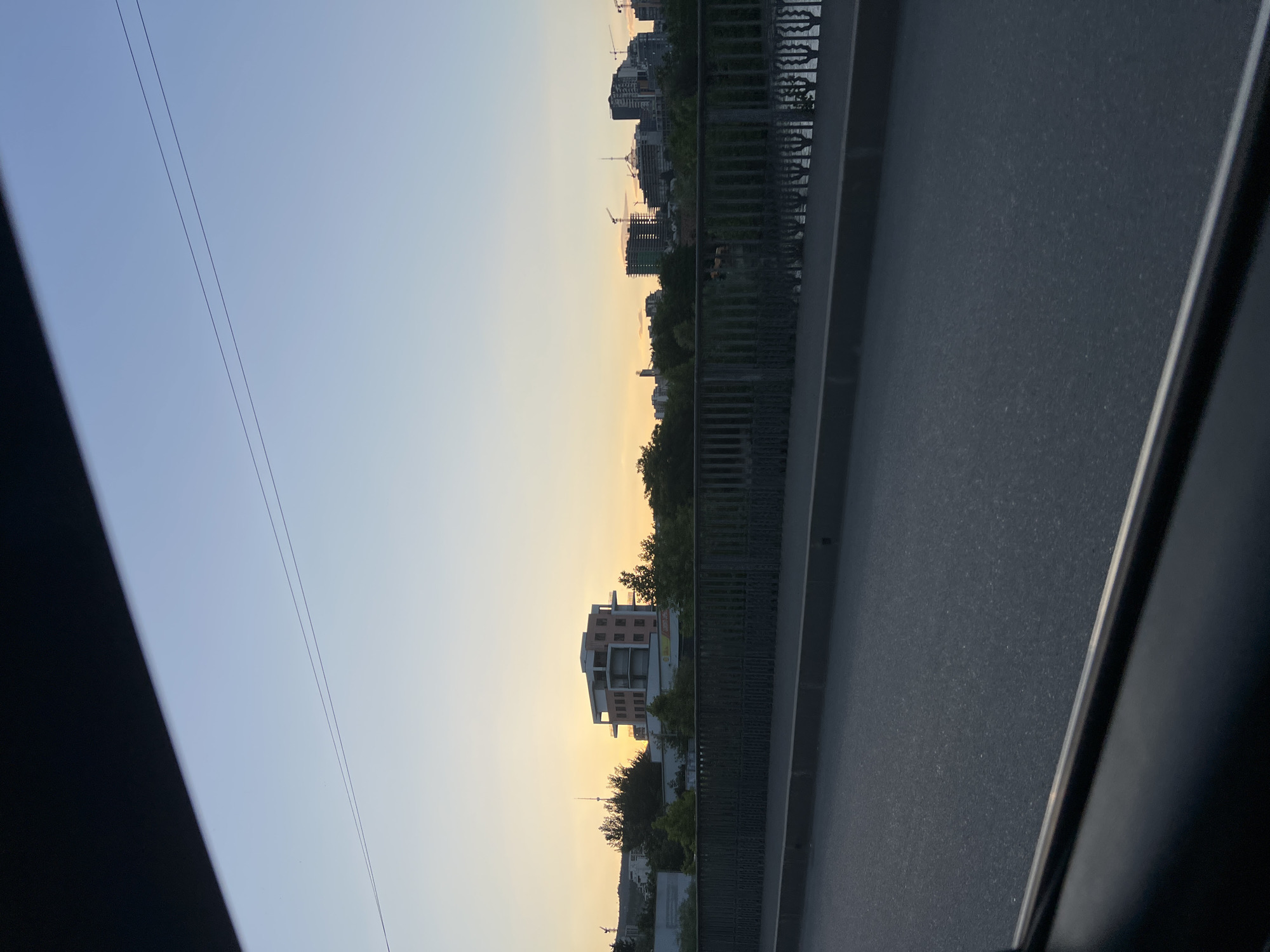 The image captures a view of the city of Tbilisi, Georgia, from what appears to be a bridge or elevated roadway. The time of day is likely sunset, given the warm glow on the horizon and the clear, light blue sky.
In the mid-ground, a multi-story building with a light-colored facade and a darker top section is prominent. It features multiple balconies and windows. Behind this building, a dense line of trees obscures much of the lower landscape. Further in the distance, a cityscape emerges, characterized by various buildings, some appearing to be under construction with cranes visible. These structures suggest a developing urban area. The presence of a body of water, likely the Mtkvari River, is implied by the reflections and the presence of a bridge railing in the foreground.
The foreground is dominated by the surface of a road, showing asphalt and a curb. A dark, shadowed area, possibly the interior of a vehicle or a structural element, frames the bottom left corner of the image. A metal railing runs along the edge of the elevated structure, with vertical supports and horizontal bars. The overall impression is of a contemporary urban environment at dusk, with a blend of older and newer architecture. No people are visible in the frame.