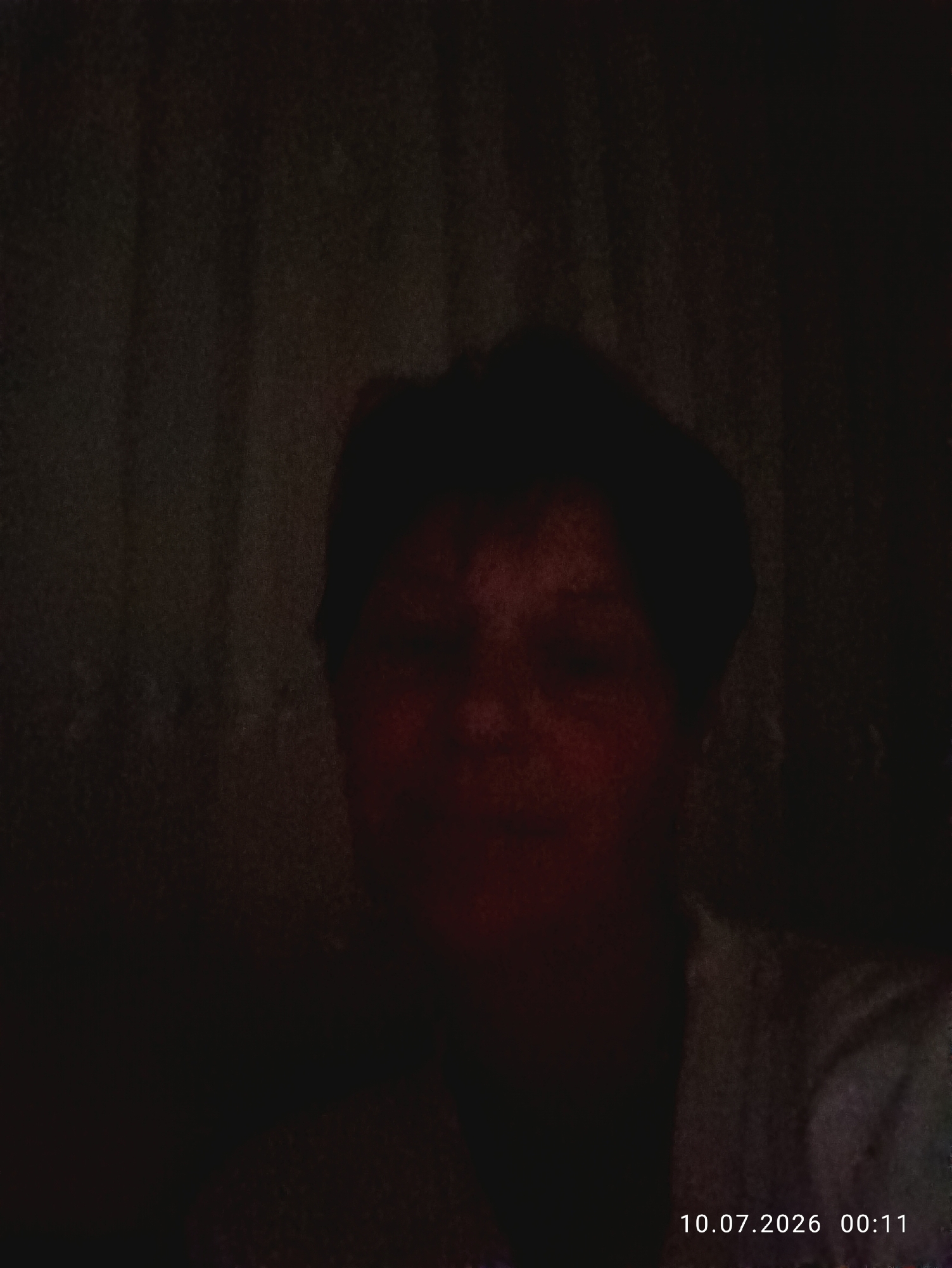 The image is a dimly lit photograph of a person's face, partially obscured by shadows. The person appears to have short, dark hair and is looking towards the camera. The background is dark and indistinct, with vertical lines suggesting curtains or blinds. A timestamp in the bottom right corner reads "10.07.2026 00:11". The image does not provide any information about the location in Nihoreni, Moldova.
