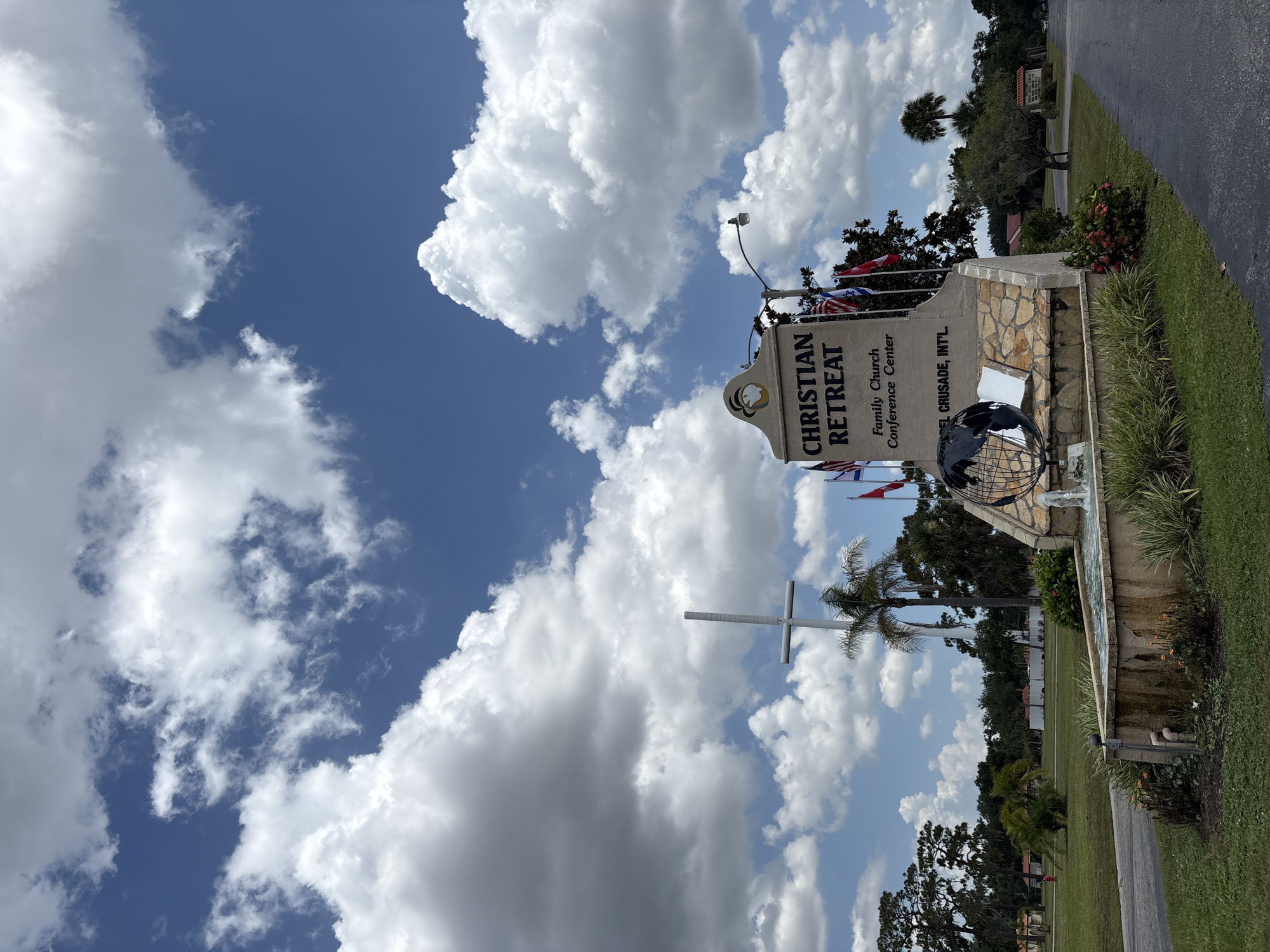 The image shows the entrance sign for "Christian Retreat Family Church Conference Center" in Bradenton, Florida, United States. The sign is made of stone and features the name of the center in large letters. Below the name, it reads "Family Church Conference Center" and "MUSEL CRUSADE, INT'L". A globe sculpture is also part of the sign. In front of the sign is a fountain with water flowing. Behind the sign, a tall white cross is visible, along with palm trees and other vegetation. Various flags are also displayed near the sign. The sky is blue with large, fluffy white clouds. The scene appears to be a sunny day, with green grass and paved pathways.