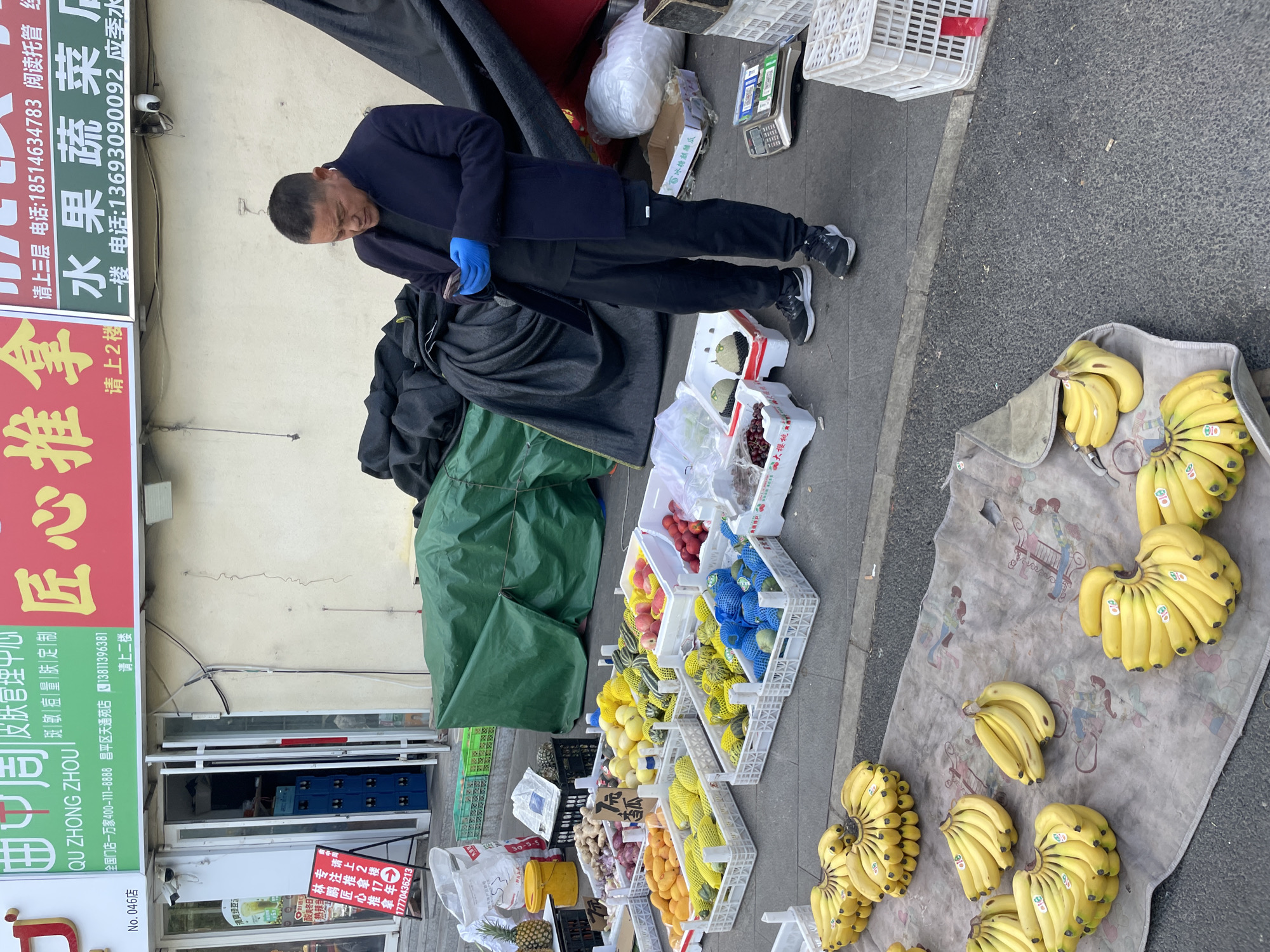 This image shows a street vendor in Changping District, China, selling fruits and vegetables. The vendor, an older Asian man, is wearing a navy blue jacket and blue gloves. He is standing near his produce, which includes bananas, oranges, mangoes, watermelons, grapes, plums, peaches, pears, pineapples, ginger, and onions displayed in white plastic crates and on a patterned tarp. Behind the vendor are black and green tarps covering other goods. A shopfront is visible, with signs for "Qu Zhong Zhou" and advertisements in Mandarin, including phone numbers.