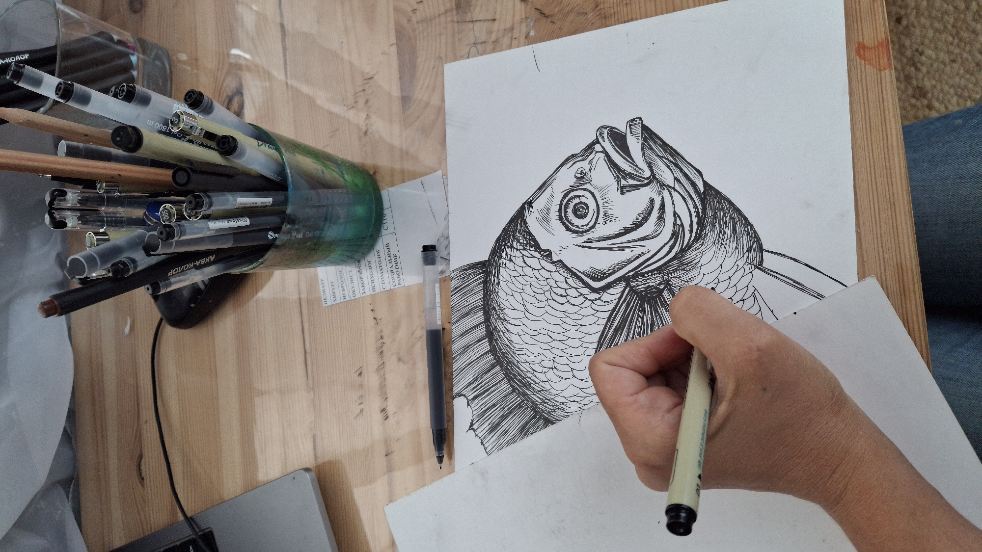 The image shows a close-up view of a hand drawing a fish on a white sheet of paper. The fish is rendered in black ink with detailed scales and fins. The hand is holding a black and white pen, actively drawing on the paper. In the background, there is a glass cup filled with various pens and pencils, as well as a laptop on a wooden table. A small piece of paper with text is visible on the table. The overall scene suggests an art or design workspace. There is no visible indication of the specific city of Kempelevo, Russia.