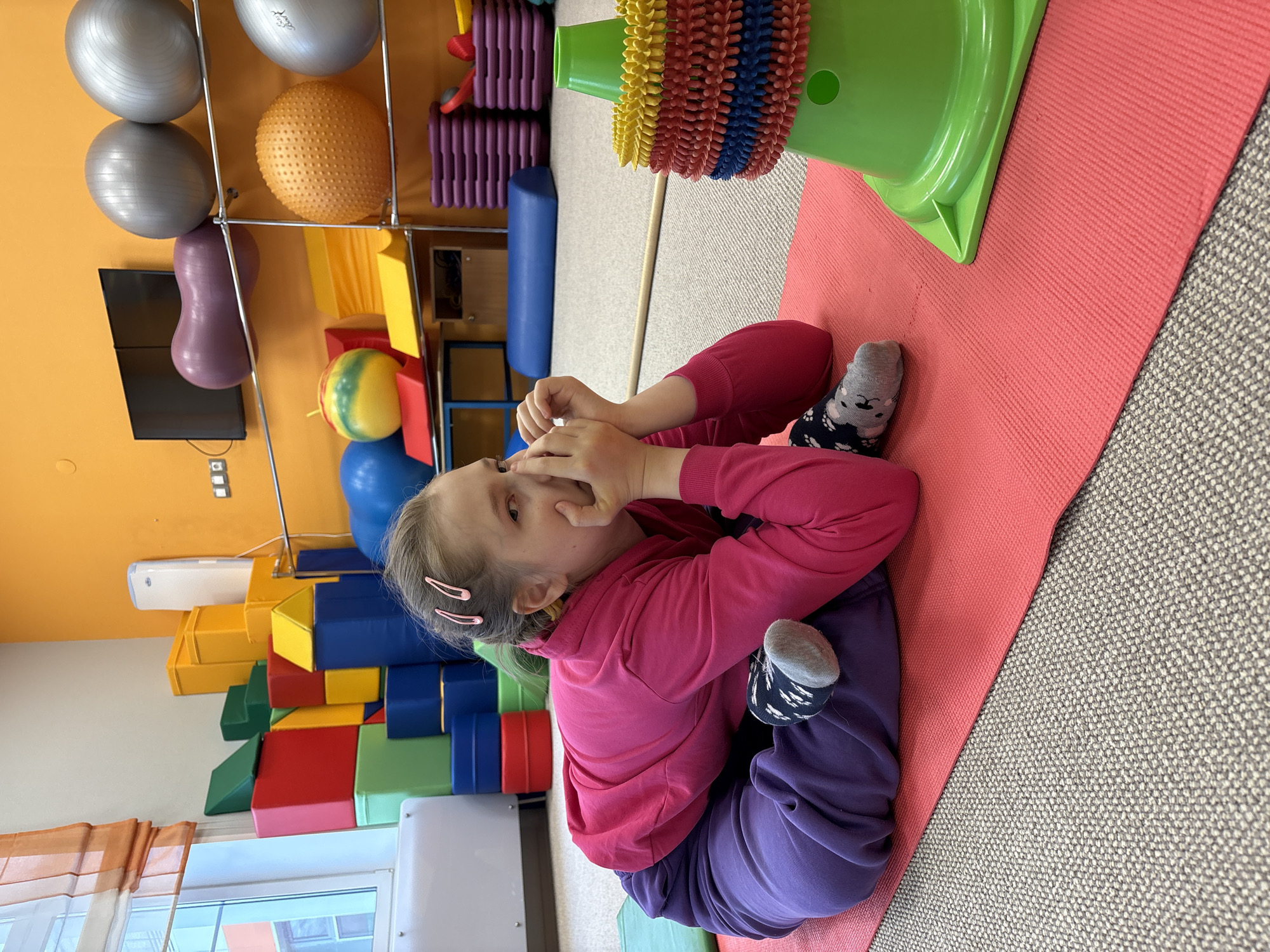 A young girl is pictured lying on a pink exercise mat in what appears to be a brightly colored therapy or activity room. She is positioned on her back with her knees bent and feet flat on the mat, wearing a pink long-sleeved top, purple sweatpants, and patterned gray socks. Her hands are brought up to her face, partially obscuring her mouth and nose, and she looks directly towards the viewer. Two pink hair clips adorn her light brown hair.
The room is filled with various exercise and sensory equipment. Against an orange wall, several exercise balls are visible, including silver, orange textured, and yellow/green striped ones, along with a purple peanut-shaped balance ball, all stored on a metal rack. A black flat-screen television is mounted on the orange wall. On the floor to the left, there is a stack of colorful soft play blocks in red, blue, yellow, and green. A large green cone-shaped object, decorated with rings of spiky, colorful sensory material (red, yellow, blue), sits on the pink mat near the girl's feet. The room has a light gray textured carpet and a window with sheer orange curtains. The setting suggests an environment for physical therapy, developmental play, or sensory integration activities for children. No visible text is present in the image.