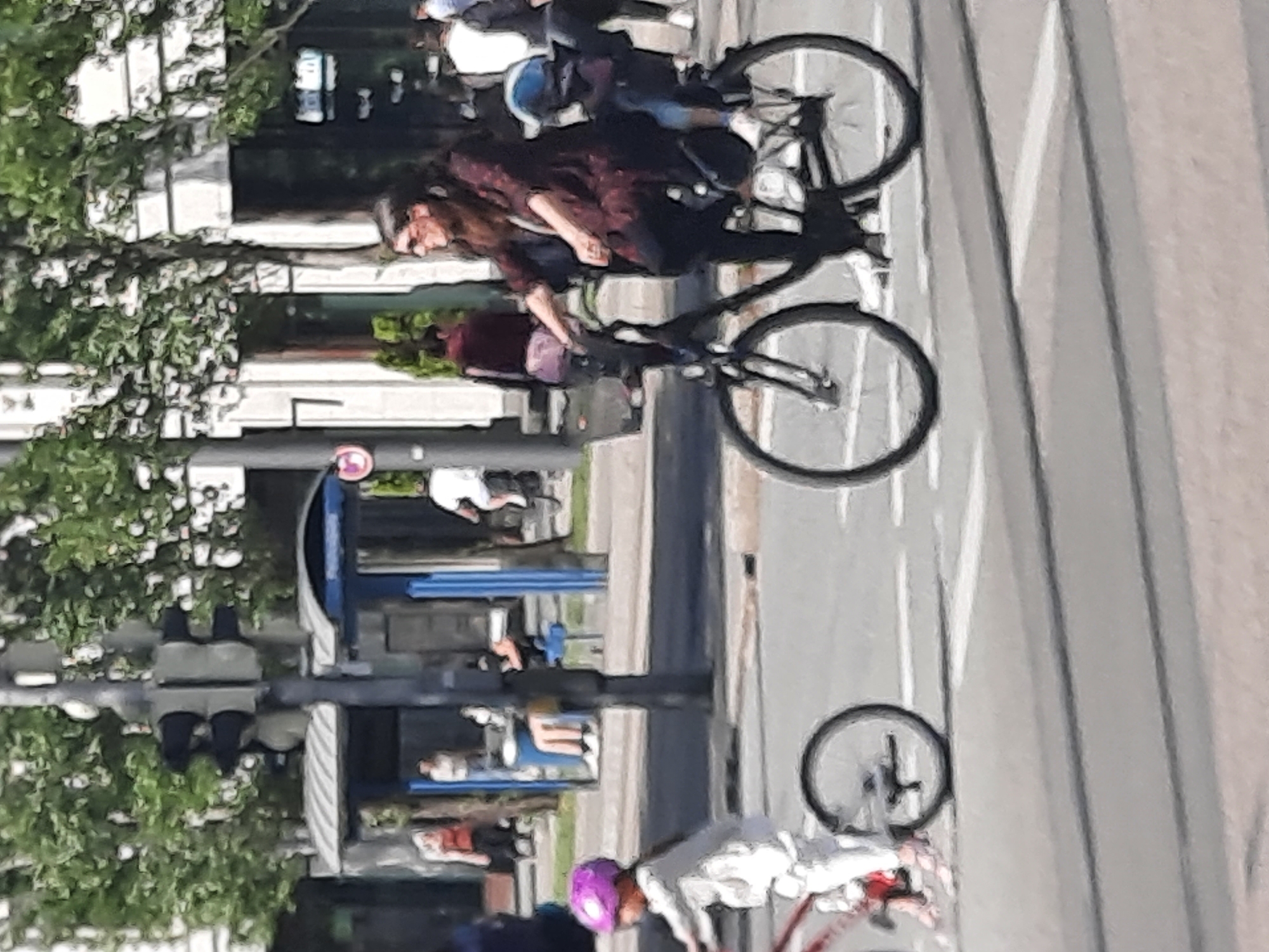 In Unknown, Unknown, a woman rides a bicycle with a child in a car seat on the back. The woman is wearing a dark patterned top and dark pants, and sunglasses. The child is wearing a blue helmet. In the foreground, a child in a white outfit and a purple helmet rides a bicycle. The background shows trees, a bus stop, and buildings. The image is taken on a street with a crosswalk.