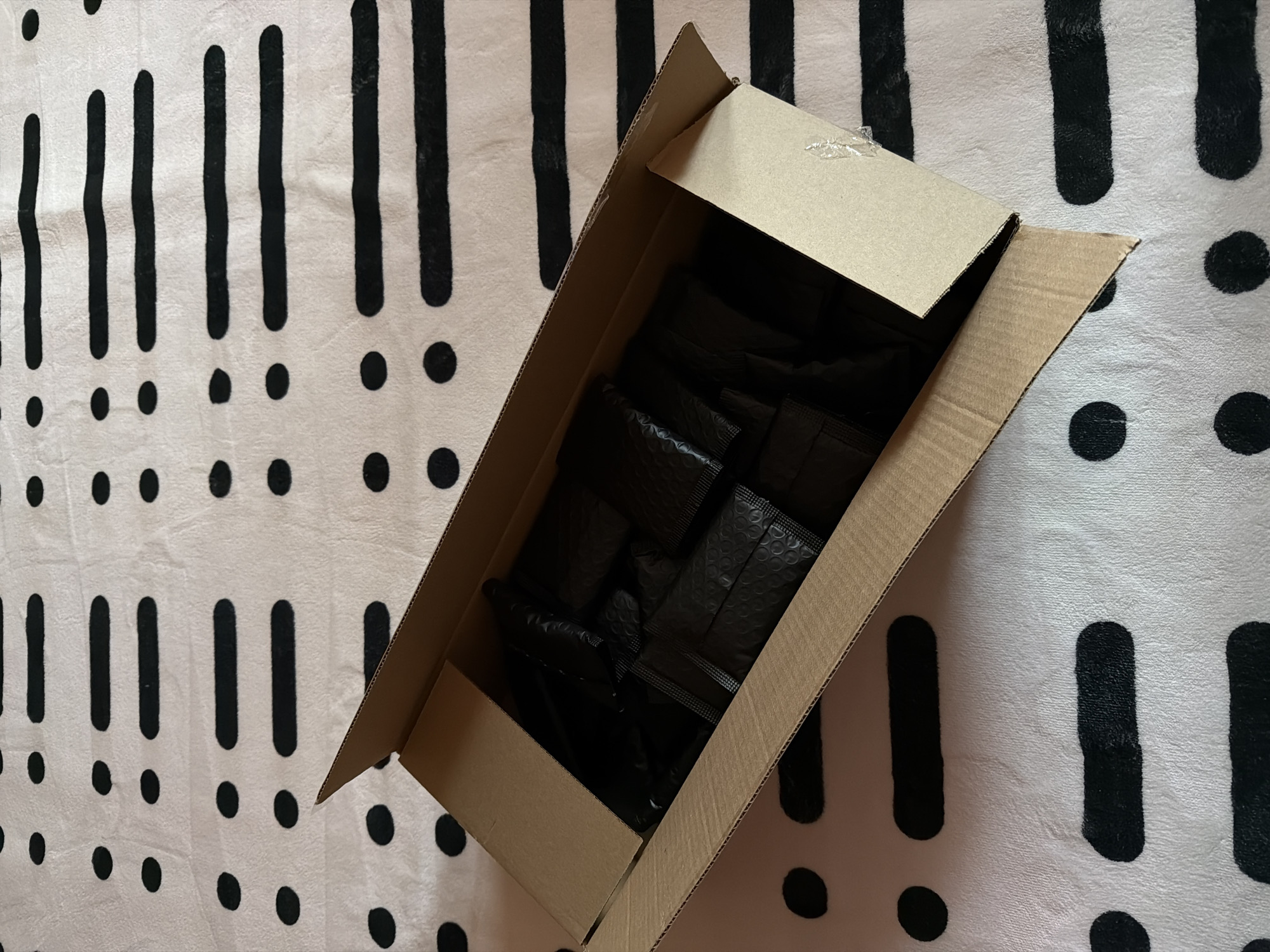 An overhead shot depicts an open cardboard box filled with multiple black padded envelopes, positioned on a patterned surface. No people are visible in the image.
The main subjects are a rectangular brown cardboard box, likely a shipping or storage container, and its contents. Inside the box are numerous small, rectangular black envelopes or bubble mailers, stacked and arranged loosely. The envelopes have a slightly textured or dimpled surface, typical of padded packaging. One side flap of the cardboard box is partially visible at the top, showing a small piece of clear adhesive tape or residue.
The box rests on a light-colored, possibly white or cream, soft fabric surface, such as a blanket or rug. This surface is adorned with a repetitive and abstract pattern of bold black exclamation marks (vertical bars with a dot below) and isolated black dots, distributed irregularly across the fabric. The lighting appears even, suggesting an indoor setting.
The image captures a static scene, possibly showing items being prepared for shipment, recently unpacked, or stored. There is no specific activity taking place beyond the arrangement of the objects.
Notable details include the contrasting black and white pattern of the background, which is quite distinct, and the uniform dark color of the envelopes, which fill the box almost entirely. The time of day and weather are not discernible. No emotions are conveyed, and no visible text appears on any of the objects or the background pattern. The location context provided is Khimki, Russia, though no specific visual cues within the image confirm this geographical detail.