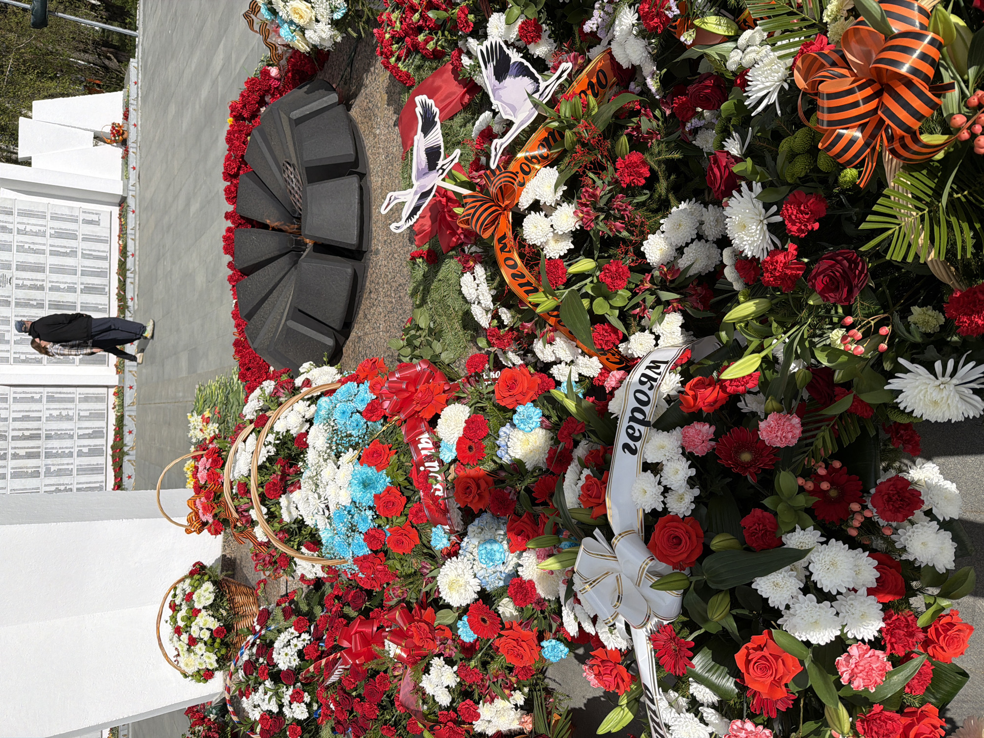 The image shows a monument in the city of Chaikovsky, Russia. The main feature of the scene is a star-shaped eternal flame surrounded by wreaths of red, white, and blue flowers. Some wreaths have ribbons with inscriptions in Russian. Also present are paper crane decorations attached to some wreaths. A person is visible in the background near a memorial wall with engraved names. The monument is set on a concrete surface. The scene is outdoors during the day with sunlight.