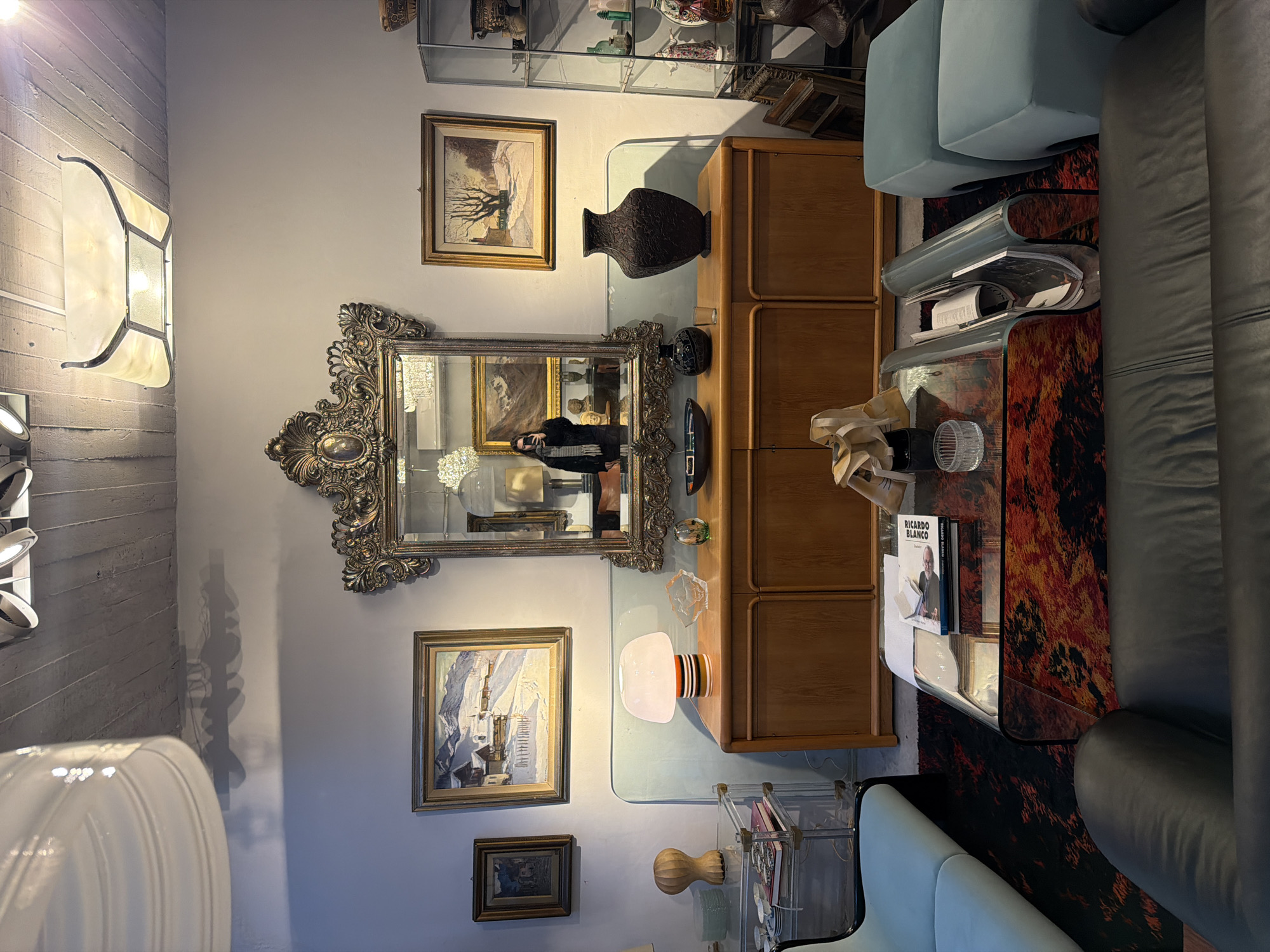 A horizontal interior shot displays a room with a mix of furniture and decor. A large, ornate mirror reflects a person taking the photograph. Above and below the mirror are framed paintings. Adjacent to the mirror and paintings is a wooden cabinet topped with a glass surface. Various decorative objects, including a dark vase and a white lamp with red and orange stripes, sit on the cabinet and glass surface. A modern glass coffee table holds books and sculptures. A blue armchair and a grey sofa frame the right side of the image. A glass shelving unit is visible near the bottom, holding decorative items. The wall is a neutral color, and the lighting appears to be a mix of natural and artificial. The location is suggested to be Buenos Aires, Argentina.