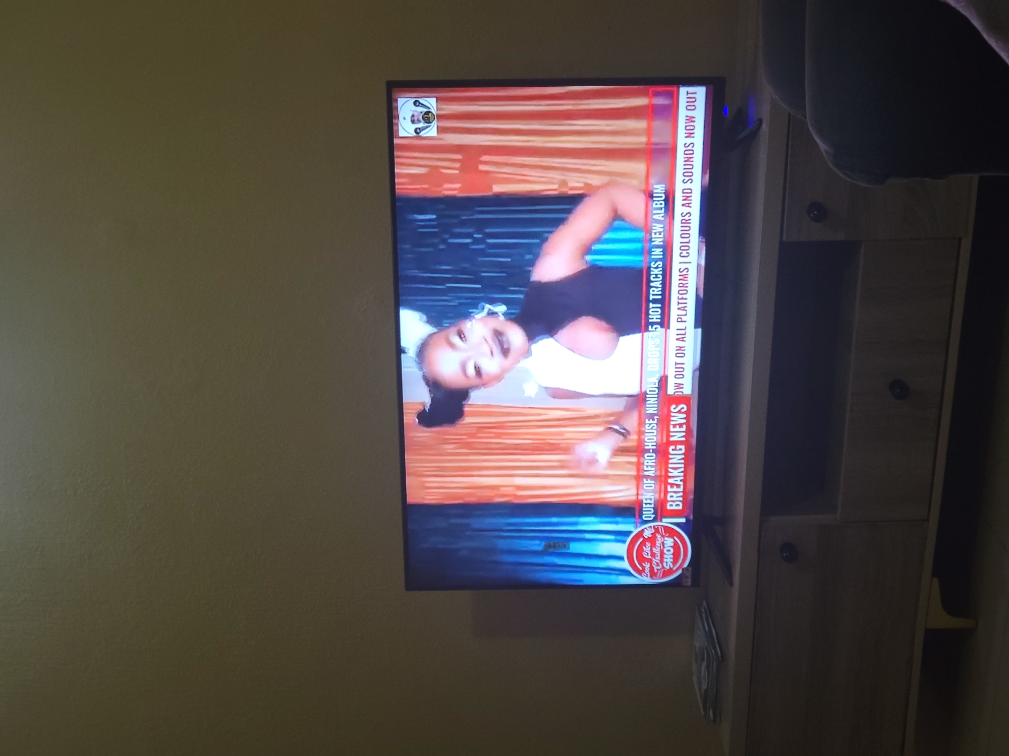 In Suleja, Nigeria, a television displays a news segment featuring a woman identified as the "Queen of Afro-House, Niniola." She is shown with dramatic makeup and a black and white outfit against a backdrop of orange and blue curtains. A lower banner on the screen reads "BREAKING NEWS" and announces that Niniola is dropping "5 HOT TRACKS IN NEW ALBUM" which is "OUT ON ALL PLATFORMS | COLOURS AND SOUNDS NOW OUT." The television is placed on a light wood entertainment stand. To the left of the television, a stack of what appear to be CDs or DVDs is visible on the stand. The room is dimly lit, with the walls painted a light brown or tan color. A dark armchair is partially visible in the lower right corner of the frame.