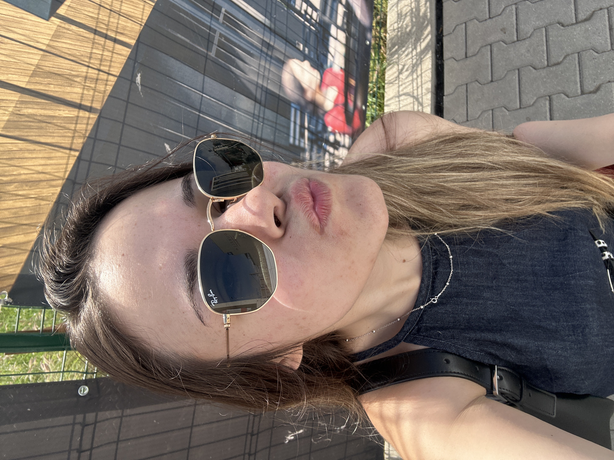 The image is a rotated, close-up selfie of a young woman taken outdoors on a sunny day in Sofia, Bulgaria. She is captured from the chest up, with her head tilted and her gaze directed slightly upwards towards the camera. Her lips are pursed into a pout. She has long, light brown hair and fair skin with visible freckles.
She is wearing square-framed sunglasses with gold-colored metal arms; the brand "Ray-Ban" is clearly visible on the right lens. Her attire includes a dark blue or grey denim top, and she wears a delicate silver-colored necklace adorned with small beads. A black leather strap, likely from a bag, is visible over her right shoulder.
The bright sunlight casts distinct shadows across the background. To her left, a wooden or wood-patterned surface is visible with strong, linear shadows. Behind her, a dark wall features what appears to be a mural or large poster with abstract shapes, including a red form, partially obscured by a fence-like structure. To her right, grey interlocked paving stones indicate an urban outdoor setting such as a sidewalk or public square. Sparse green foliage is visible along the edges of the frame. The overall impression is a casual, bright moment captured outdoors.