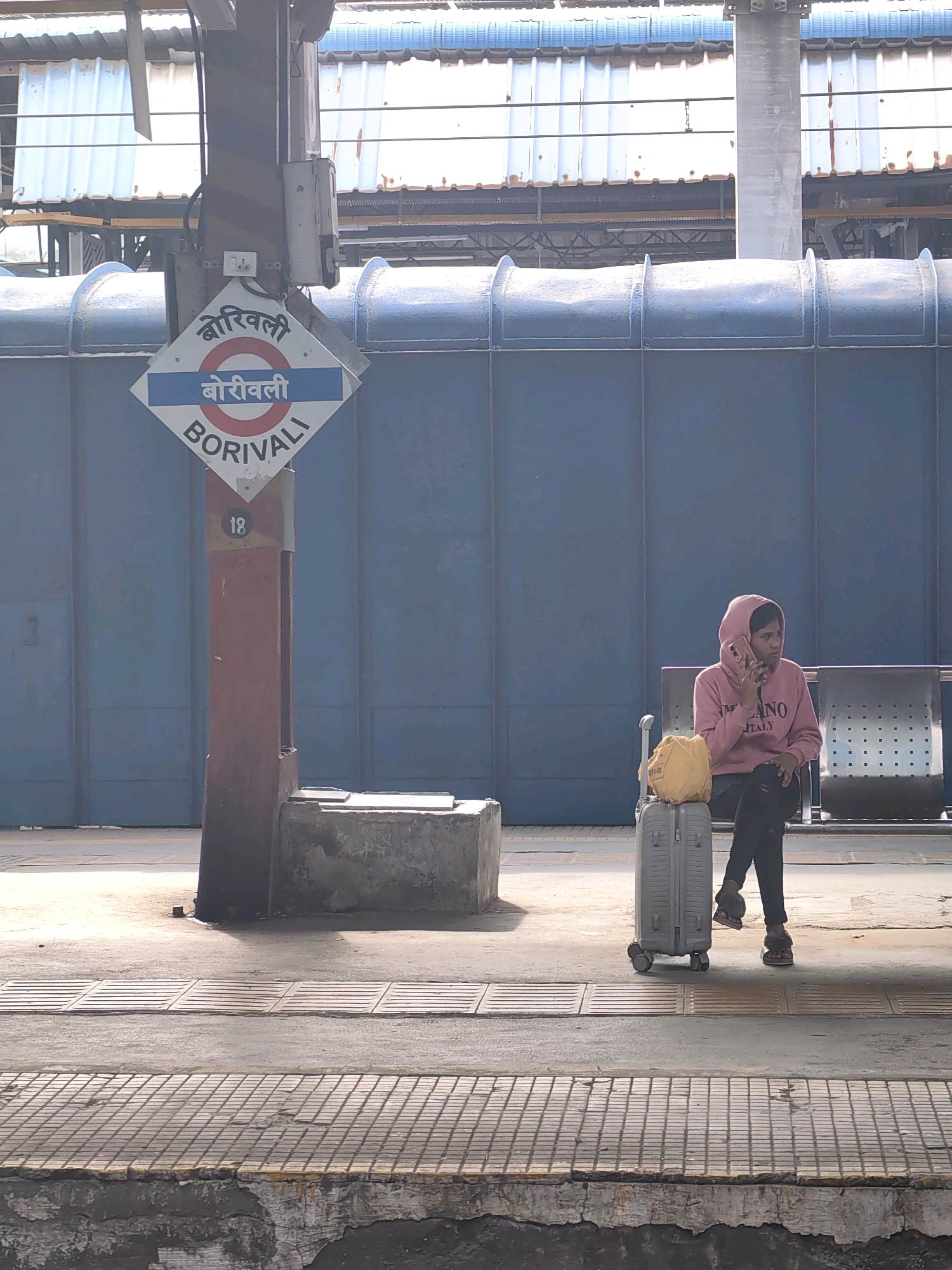 A young woman in a pink hooded sweatshirt with the words "MILANO ITALY" is sitting on a bench at a train station, talking on a pink mobile phone. She is wearing black pants and fluffy slippers. Beside her is a grey suitcase with a yellow bag on top. Behind her, a blue metal wall stretches across the background. In the foreground, a signpost with the name "BORIVALI" written in Hindi and English indicates the location of the Borivali railway station, a major suburban railway station in the city of Mumbai, India. The platform is made of concrete and tiles, with a visible drain cover. The scene is outdoors, under a covered structure.