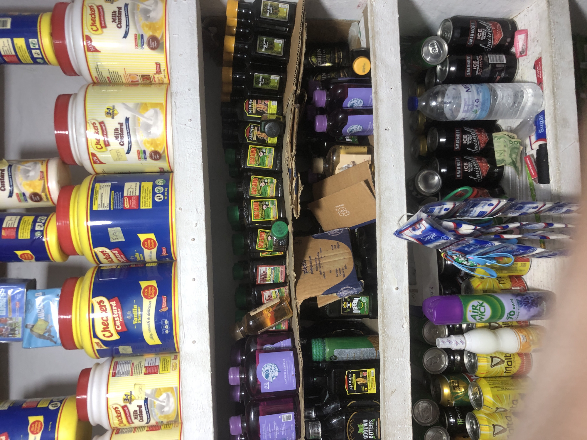 The image shows a cluttered shelf in what appears to be a small store or stall. The shelves are filled with various products, including bottles of what looks like liqueur and medicinal tonics, cans of beverages, and containers of custard powder. In the foreground, there are also some packets of sugar and currency. The overall impression is one of a busy retail environment with a wide assortment of goods. The scene is brightly lit, and the shelves are made of wood. There are no people visible in the image.