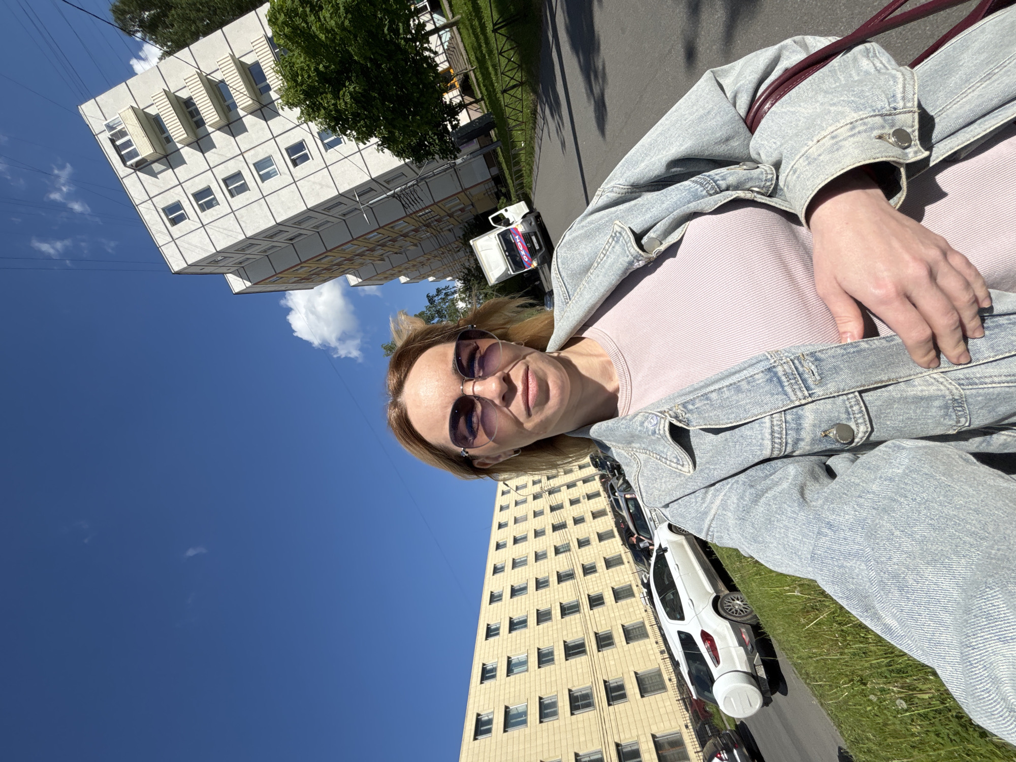 The image captures a woman taking a selfie on a bright, sunny day in an urban or suburban environment, rotated 90 degrees clockwise.
In the foreground, a woman with shoulder-length blonde or light brown hair wears sunglasses with purple-tinted lenses, looking towards the camera with a neutral or slightly smiling expression. She is dressed in a light pink ribbed top layered with a light blue denim jacket, and her right hand is visible, resting near her waist.
The background is dominated by a clear, vibrant blue sky with a few sparse white clouds, indicating a fine, daytime setting. To the left (top of the rotated image), a multi-story white or light grey building features a grid-like facade with numerous rectangular windows and several distinctive projecting balcony-like structures. Further down the street, another tall, light yellow or beige multi-story building with many windows provides a receding backdrop.
Along the street, several cars are parked, including a white SUV or hatchback with a spare tire mounted on its rear. A small white delivery van is partially visible behind the woman, prominently displaying the word "OZON" in blue lettering, identifying it as a vehicle for the Russian e-commerce company and confirming the implied location in Russia. Green grass and trees line the street, adding natural elements to the built environment.
The scene depicts a casual outdoor moment under clear skies, likely in a residential or mixed-use area.