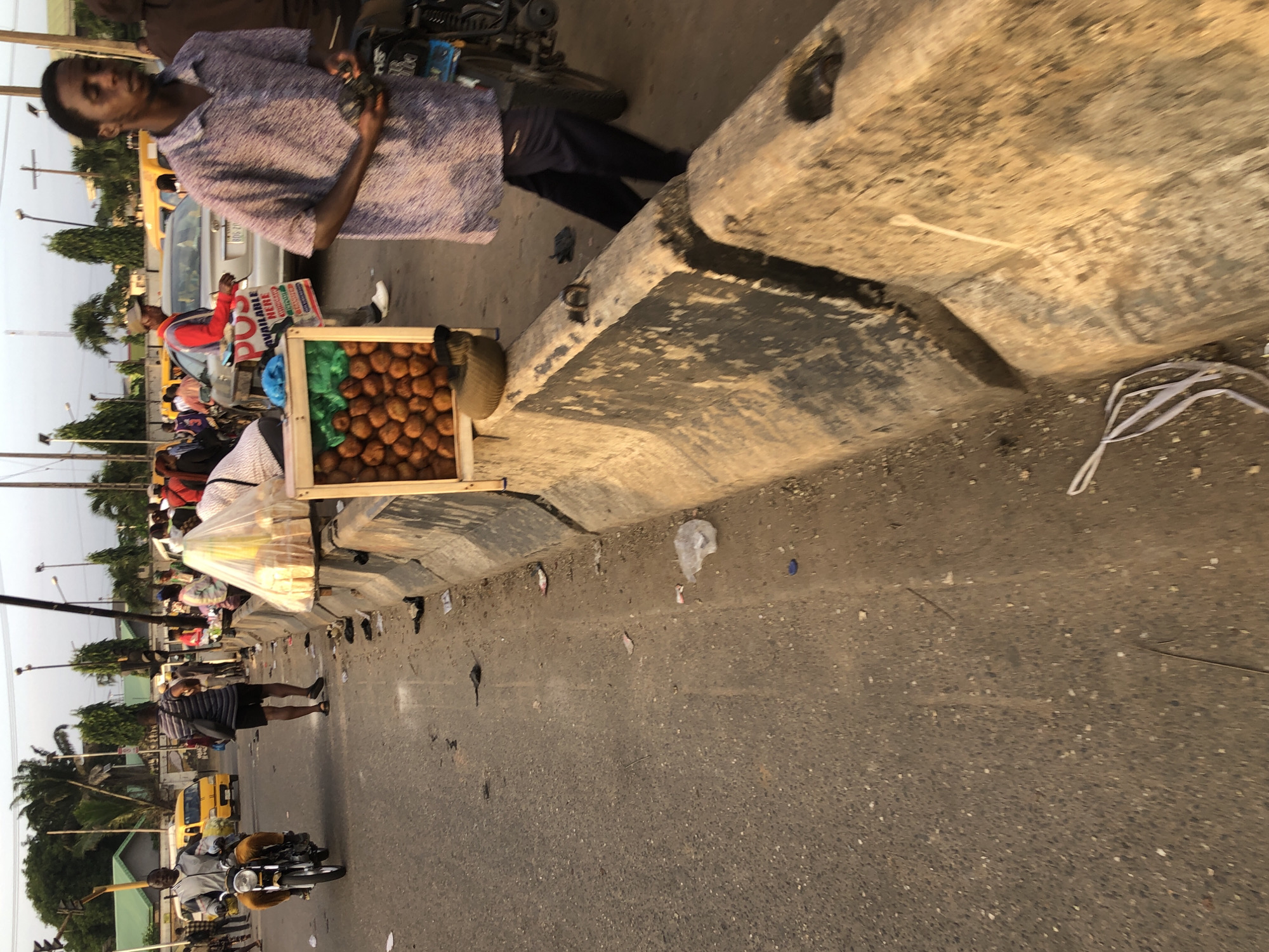 This is a medium shot, taken at eye level, of a street scene in Ikeja, Nigeria. The image captures a busy street with pedestrians and vehicles. In the foreground, a man wearing a purple patterned shirt and dark pants is standing on the right side of the frame, holding what appears to be a black device. To his left, a wooden stand is filled with small, round, brown objects, possibly food items. This stand is placed on top of a concrete barrier. Further down the street, other pedestrians are visible, some walking and others standing. There are also vehicles, including a yellow taxi in the background and a motorcycle on the left side of the frame with a rider. The street is paved and shows signs of wear and tear, with debris scattered on the ground. The environment appears to be an urban setting with buildings and trees visible in the distance. The lighting suggests it is either early morning or late afternoon.