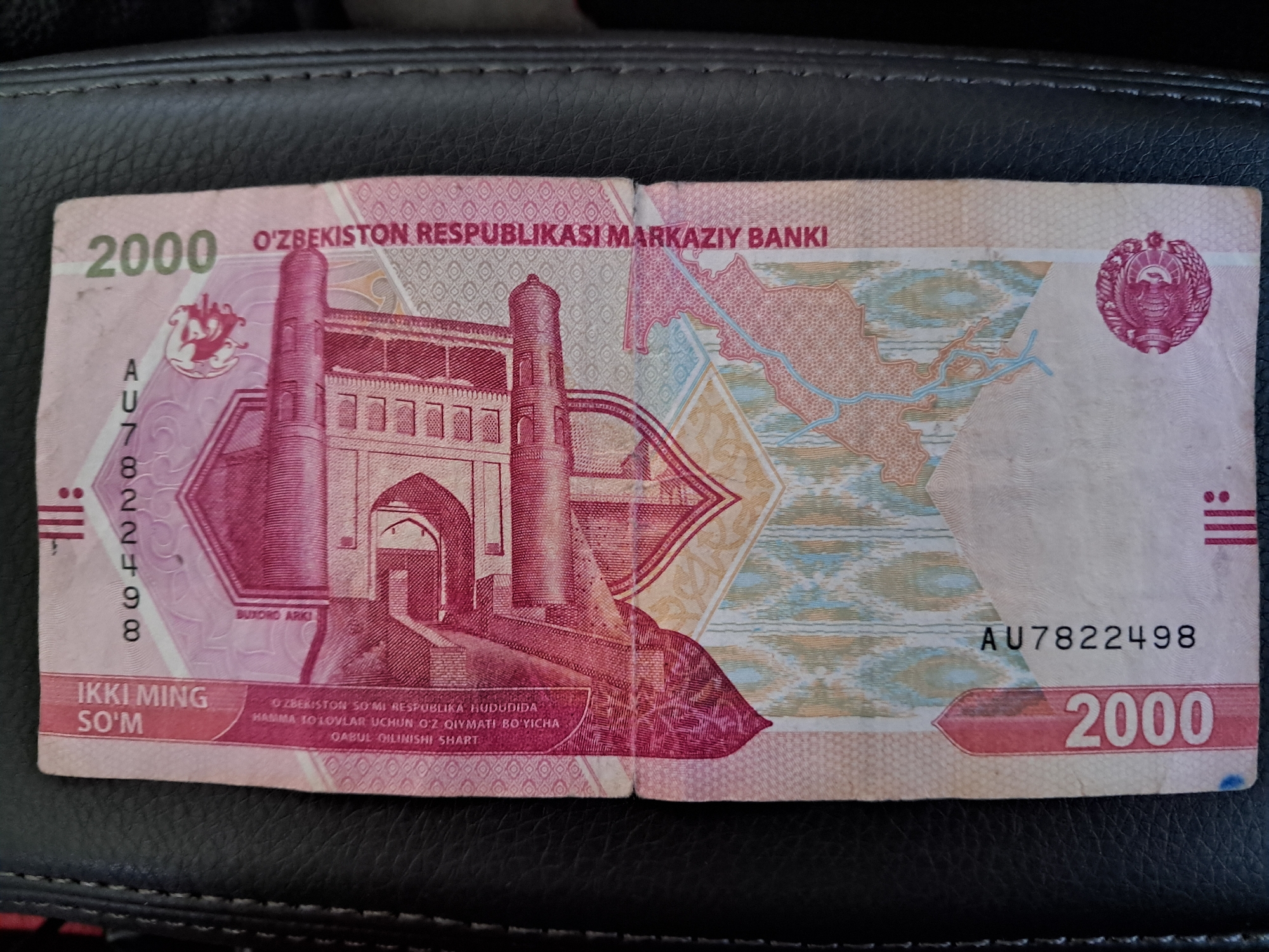 The image shows a close-up, overhead view of a two-thousand Uzbekistani Som banknote. The banknote is predominantly pink and features intricate designs, including what appears to be a building or monument in the center. The text "IKKI MING SO'M" is visible on the left side, indicating "Two Thousand Som." On the right, the text "O'ZBEKISTON RESPUBLIKASI MARKAZIY BANKI" is printed vertically, meaning "Central Bank of the Republic of Uzbekistan." The serial number "AU7822498" is present on the lower left side, and "2000" is repeated on both the left and right edges. The banknote is laid flat on a textured black surface, likely leather. There are no people or indications of the city of Bagish, Uzbekistan.