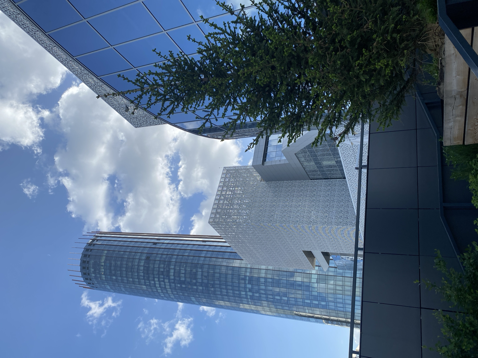 The image depicts a low-angle view of several modern buildings in Yekaterinburg, Russia, set against a bright blue sky with scattered white clouds. A tall, cylindrical skyscraper with a gridded facade dominates the left side of the frame. Adjacent to it is a cube-shaped structure with a patterned, possibly perforated, outer layer. To the upper right, a building with a reflective glass facade and decorative trim is partially visible. A tree with dense green foliage partially obscures the view in the upper right corner. The architecture suggests a contemporary urban environment.