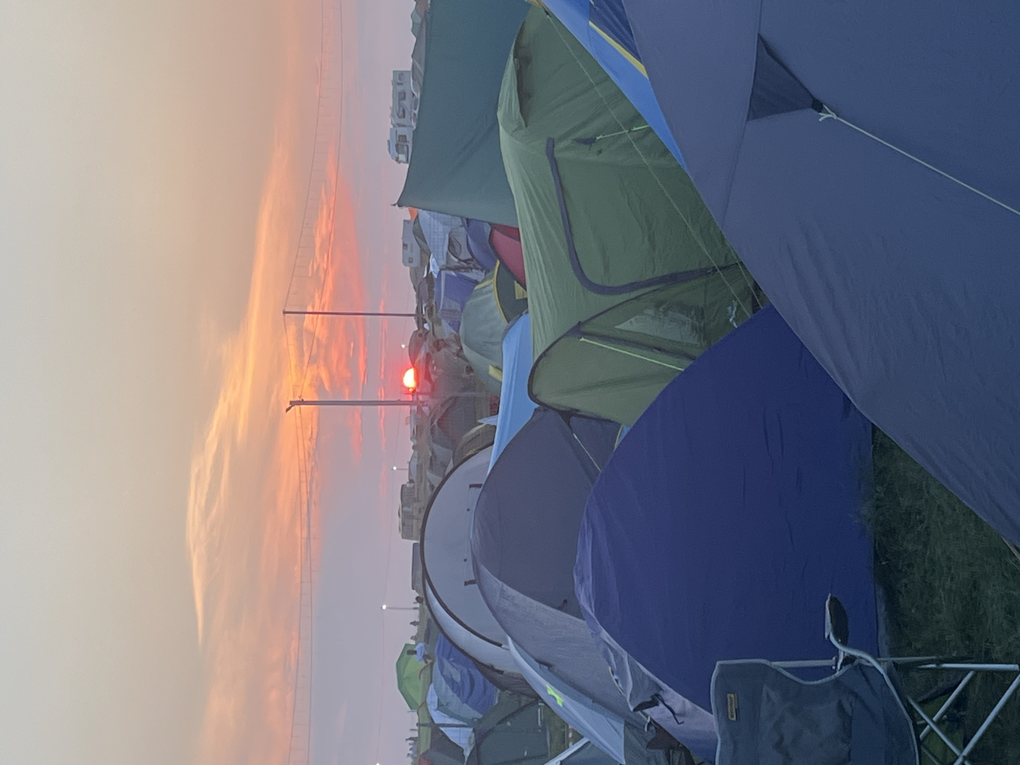 The image depicts a campsite at sunrise, with a sky ablaze with orange and pink hues. Numerous tents of various colors, primarily blue, green, and gray, are pitched closely together, forming a dense field of temporary shelters. In the background, faint silhouettes of people can be seen scattered amongst the tents, and a few recreational vehicles are visible. A large, horizontal structure, possibly a bridge or a power line support, spans across the horizon. The sun, a bright orange orb, is just beginning to rise, partially obscured by clouds and the tents. The ground appears to be covered in grass, suggesting an outdoor location. The overall impression is one of a festival or a large gathering, likely at the beginning of a day.