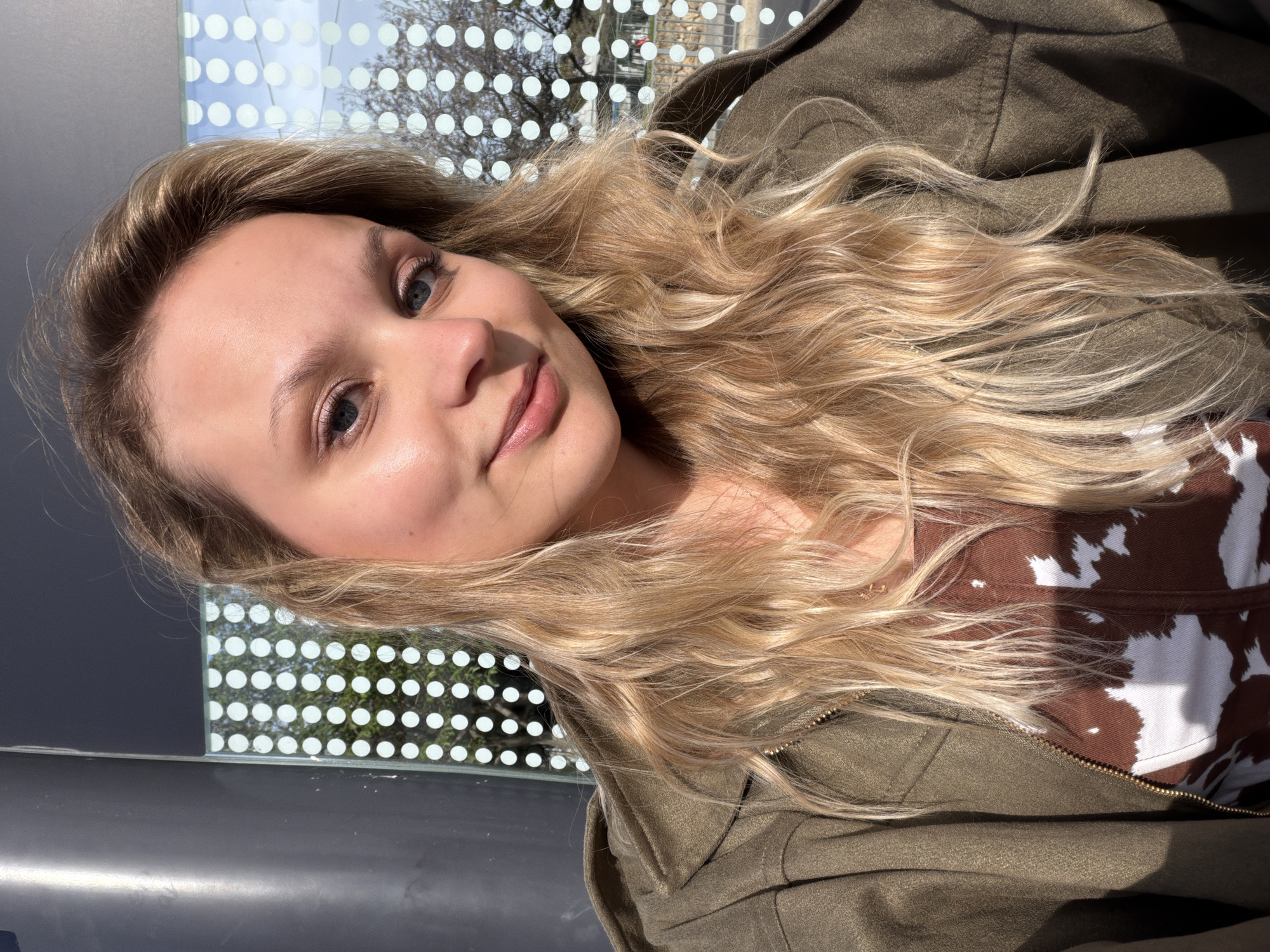 A young woman with long, wavy blonde hair is captured in a portrait-oriented, eye-level shot. She looks directly at the viewer with a soft, friendly smile, her face brightly lit by sunlight. Her blue eyes are subtly enhanced with light makeup, as are her lips.
Her blonde hair, featuring natural waves and various shades, frames her face and flows over her shoulders. She is wearing an olive-green hooded jacket, partially unzipped, which reveals a brown and white cow-print top underneath.
The background features a modern glass partition or window adorned with a pattern of evenly spaced white dots. Through this translucent screen, blurred trees with bare branches and a bright blue sky are visible, indicating an outdoor setting bathed in sunshine. Below the dotted window, a dark, reflective surface is present.
The image conveys a casual and relaxed atmosphere, likely a selfie or close-up portrait taken on a sunny day in Tel-Aviv, Israel. There is no visible text in the media file.
